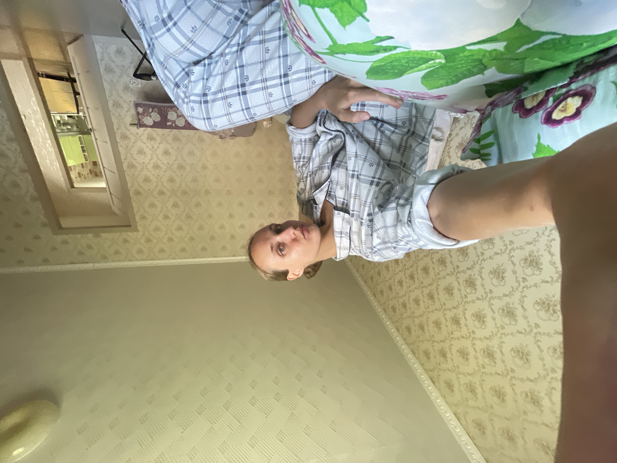 A low-angle, wide shot captures a woman in plaid pajamas looking downwards with her left arm extended towards the camera, as if taking a selfie. She is seated on a bed with a floral-patterned pillow in the foreground. The room has patterned wallpaper and a textured ceiling. In the background, a doorway leads to another room with kitchen cabinets. The lighting suggests it is daytime. The location is identified as Sharkan, Russia.