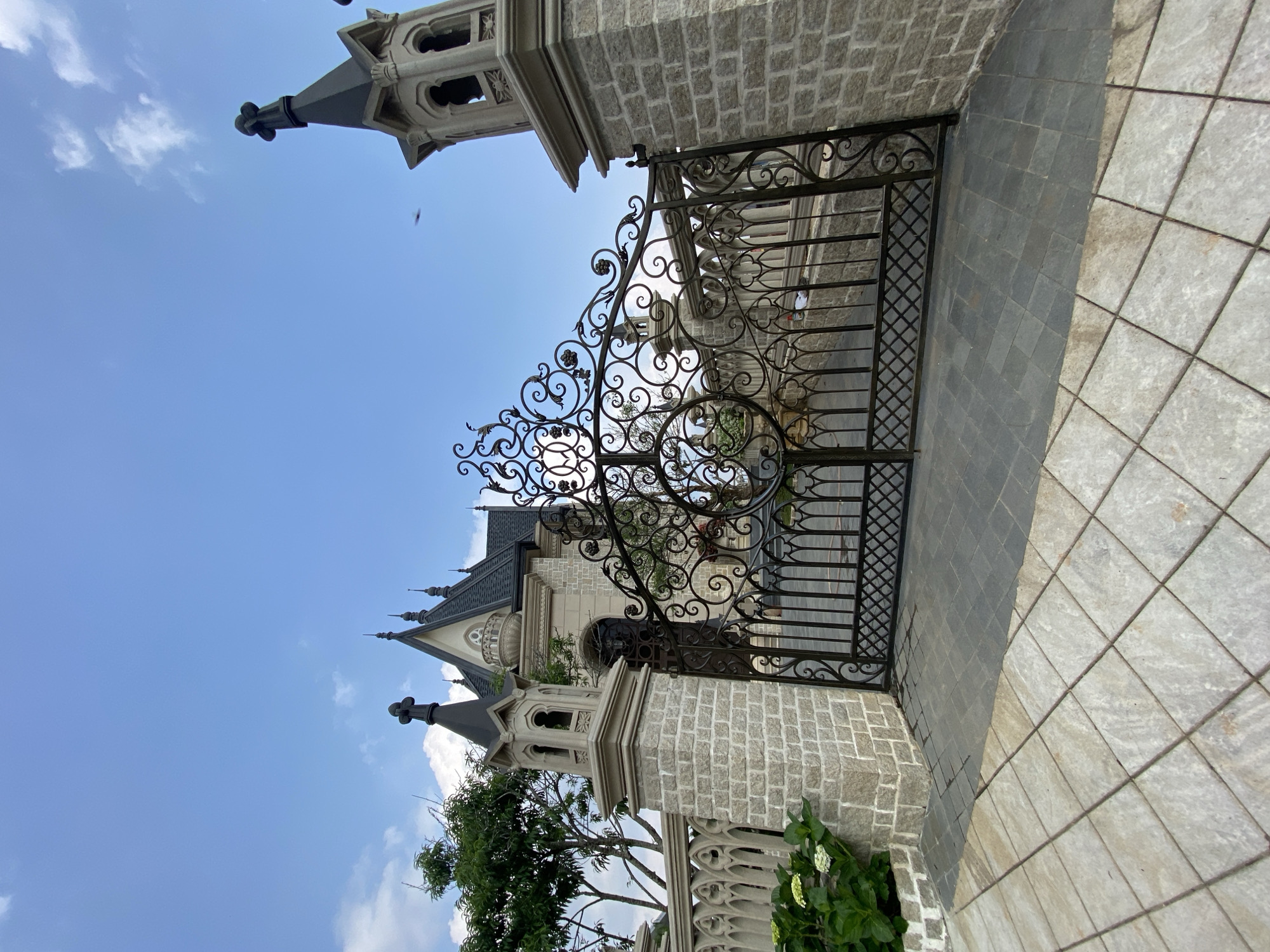 Exterior shot of the Domaine de Marie Church gate. The gate is made of dark metal and has ornate designs. The building behind the gate is constructed from light gray brick. There is a gray and white stone walkway in front of the gate. The sky is blue with some white clouds. There is a tree and some green plants near the gate.