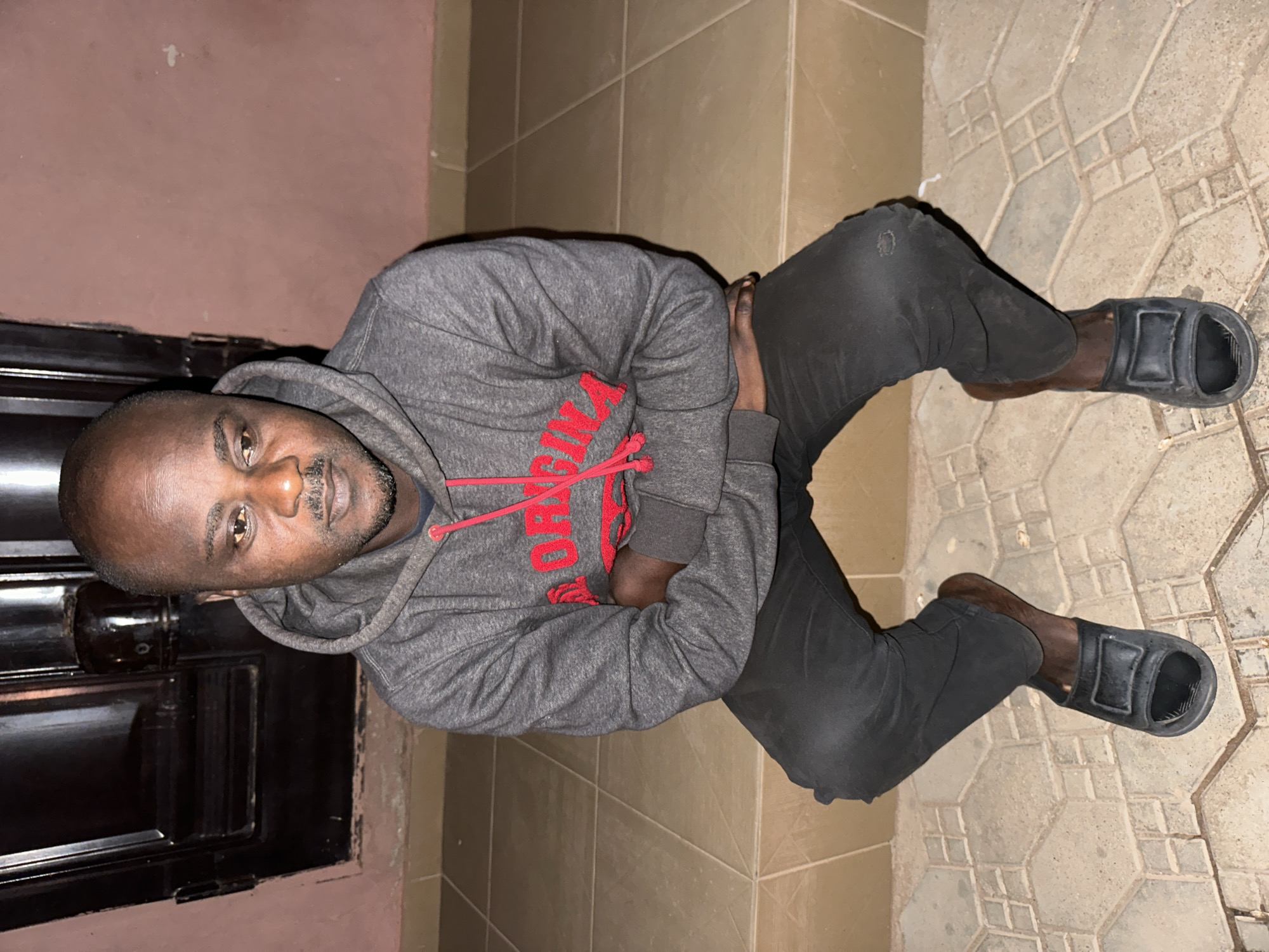 A man with dark skin and a shaved head, wearing a grey hooded sweatshirt with red lettering and black pants, sits on a tiled step outdoors. His arms are crossed, and he is looking down and to his left. The background includes a dark brown door and a pinkish wall. The ground is covered in square tiles with a geometric pattern. The scene appears to be in a residential area in Narayi, Nigeria.