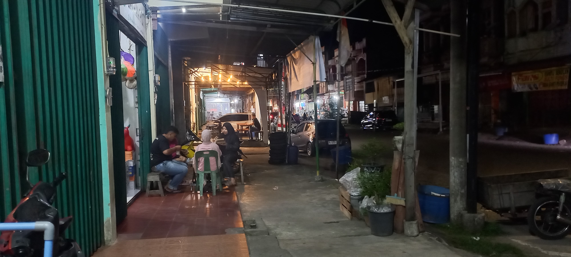 In Tapak Tuan, Indonesia, at night, a narrow street scene unfolds under artificial lighting. A green corrugated metal gate forms the left boundary of the frame, with a motorcycle parked in front of it. To the right of the gate, an open storefront reveals a space where several individuals are seated at low tables and on small stools. Three people are visible in this area: a man in a black t-shirt and jeans, a woman in a pink hijab and a black veil, and another woman in a black hijab. They appear to be engaged in conversation or eating.
Further down the street, the walkway continues under a covered structure supported by metal poles. Stacks of tires are visible on the right side of the walkway. Several cars are parked or driving in the background, including a dark sedan and a silver hatchback. Buildings line the right side of the street, with illuminated storefronts and advertisements. The ground is a mix of concrete and tiled surfaces. In the distance, more buildings and streetlights are visible, indicating an urban environment. Two blue barrels are present on the right side of the street, one near the tires and another further down. A broom leans against some wooden planks on the right. The overall impression is of a busy, commercial street at night.