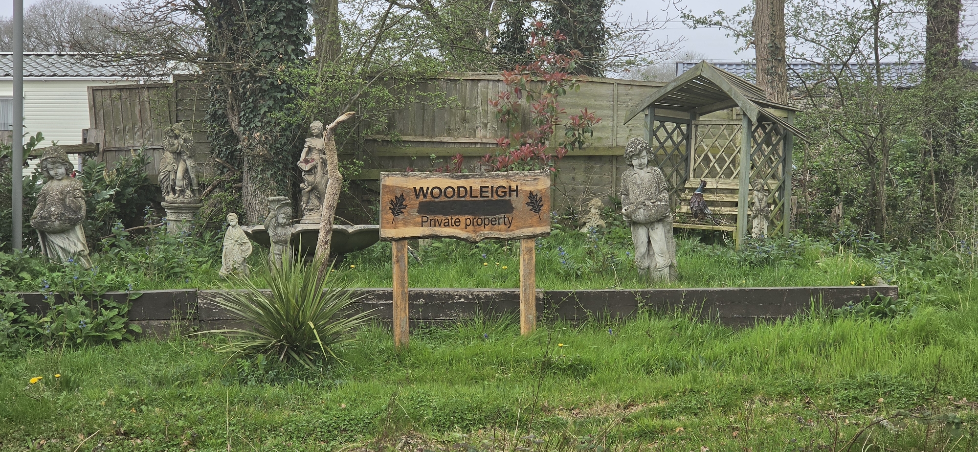 The image depicts a garden in Reigate and Banstead, United Kingdom, marked by a wooden sign reading "WOODLEIGH Private Property". Several weathered stone statues of figures are distributed amongst dense green vegetation. A weathered wooden pergola stands to the right. The garden is enclosed by a wooden fence with vines and some red-leaved foliage growing upon it. A grey, overcast sky is visible. A caravan or mobile home is visible behind the fence on the left. A raised wooden planter separates the foreground from the garden.