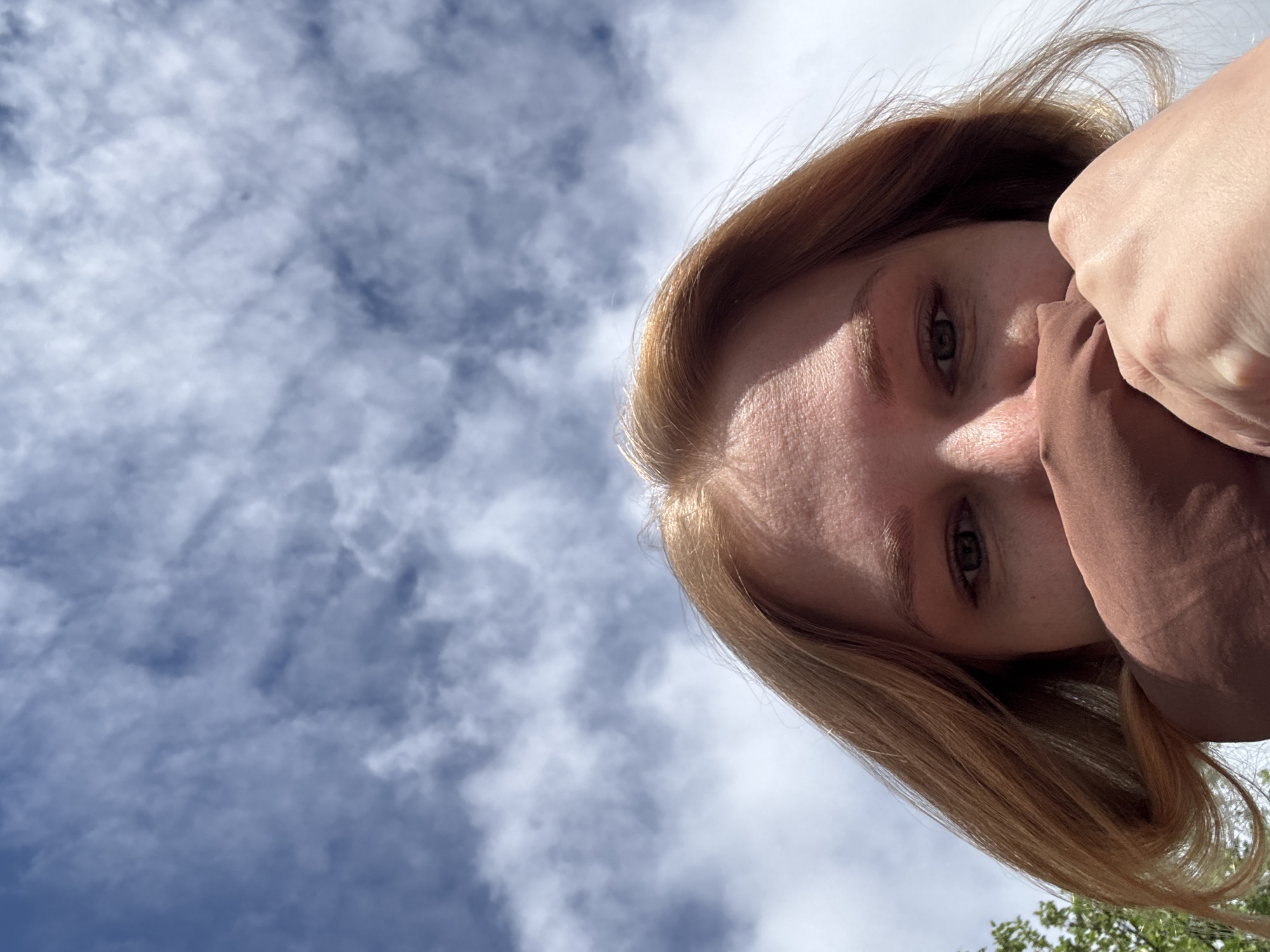 An upward-tilted shot of a woman's face partially obscured by a brown cloth held to her nose and mouth. Her hair is light brown, and the sky with blue sky and clouds is behind and to the left of her head. Greenery and tree branches are visible in the lower right corner. No information indicates this image was taken in Rusanovo, Russia.