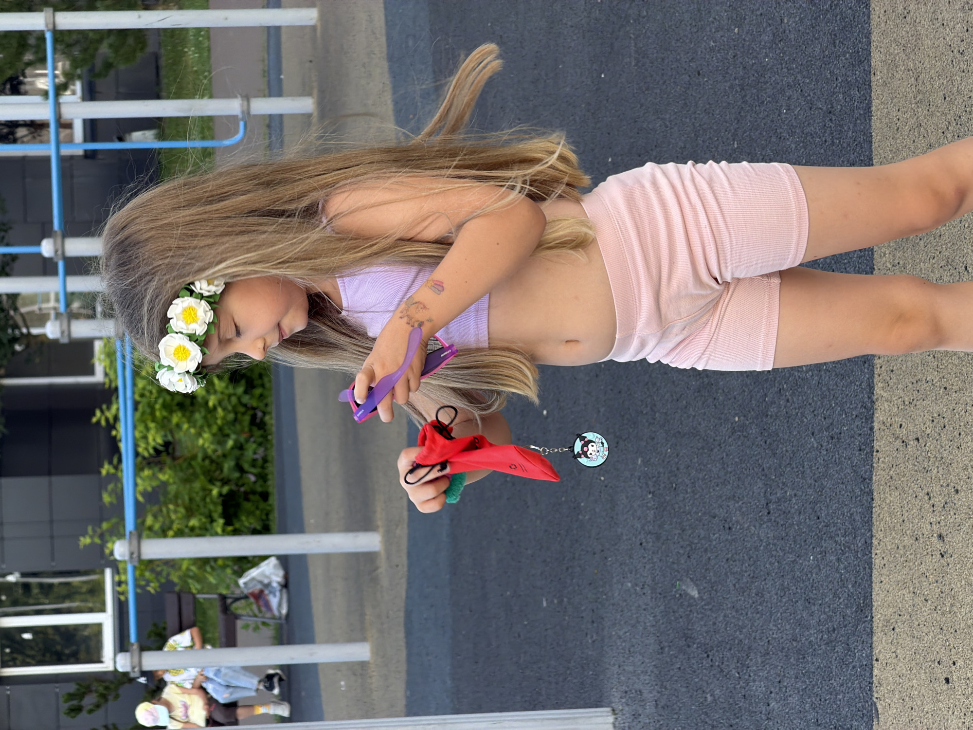 The media file captures a young girl with long blonde hair, adorned with a floral wreath, standing outdoors on a paved surface. She is wearing a light purple crop top and pink ribbed shorts. In her hands, she holds a small red pouch with a black drawstring and a keychain featuring a cartoon character. Her gaze is directed downwards, and her expression suggests focus or contemplation.
The setting appears to be a playground or park area, with a metal frame structure and greenery visible in the background. The ground is a mix of dark grey asphalt and lighter colored pavement. The lighting suggests it is daytime, possibly late afternoon, with mild weather conditions. Other people are visible in the distance, also appearing to be in an outdoor recreational space.
The image is oriented vertically, with the girl on the right side of the frame. The overall mood is casual and playful, fitting for a child in an outdoor environment. There is no visible text in the image.