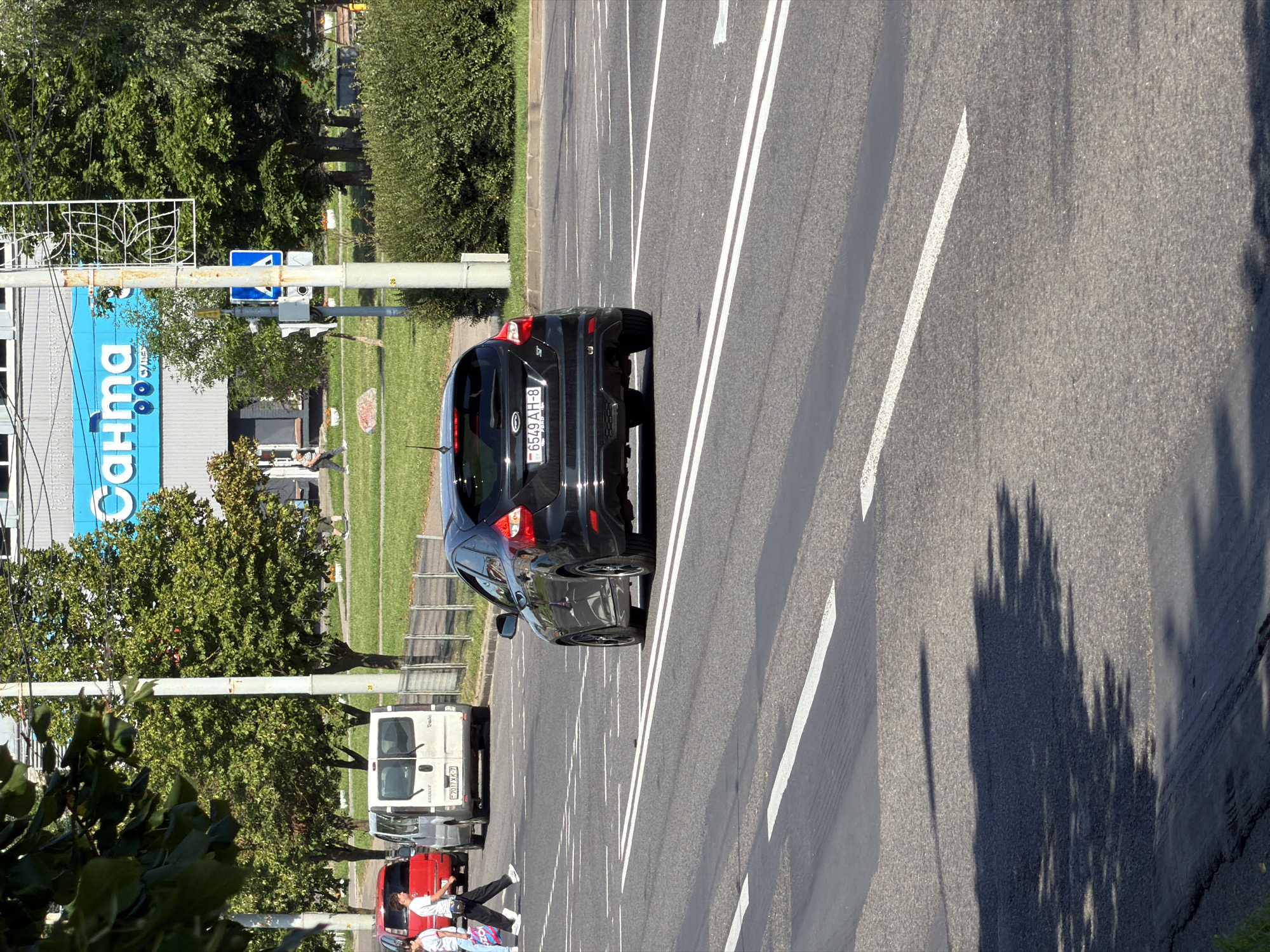 The image captures a street scene in Minsk, Belarus, on a sunny day. In the foreground, a black Ford Focus is driving on a multi-lane road with clear white road markings. Pedestrians are visible crossing the road on a designated crosswalk, and a white van and a red car are parked or driving on the side. In the background, a building with a large blue sign reading "Сантa" (Santa) is visible, flanked by trees and utility poles. The environment appears to be an urban setting with a well-maintained road and sidewalks.