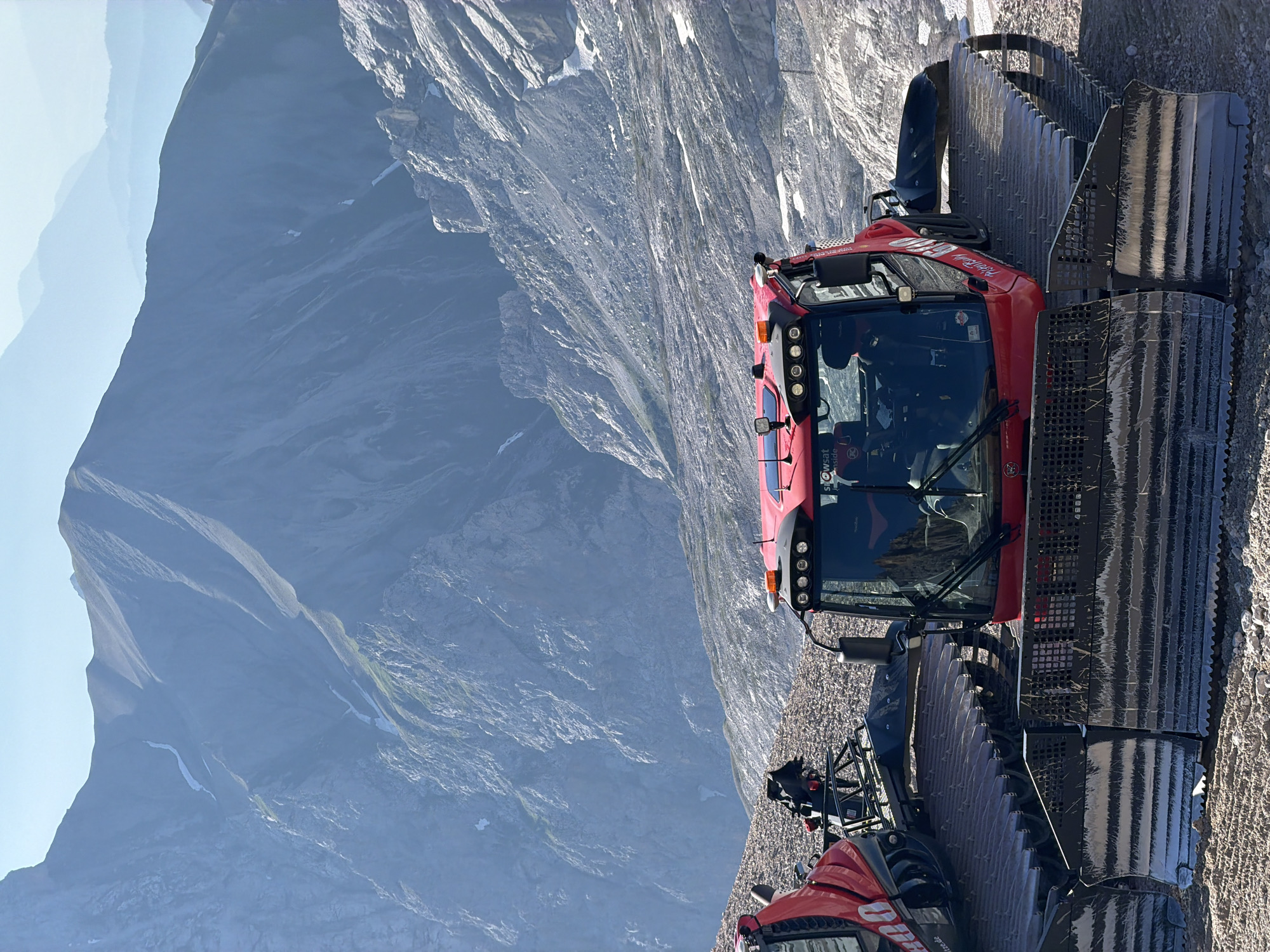 This is a medium shot of two red snow groomers parked on a rocky, mountainous terrain, likely on Zugspitze, the highest peak in Germany near Garmisch-Partenkirchen. The snow groomers are large, tracked vehicles with robust blades at the front. The one in the foreground is largely visible, showing its cab with windows and lights, and its wide tracks. The second snow groomer is partially visible to the left. In the background, layers of hazy blue mountains stretch out under a clear sky, with patches of snow visible on their slopes. The overall scene depicts a high-altitude environment, possibly at a ski resort during the off-season or in preparation for winter.