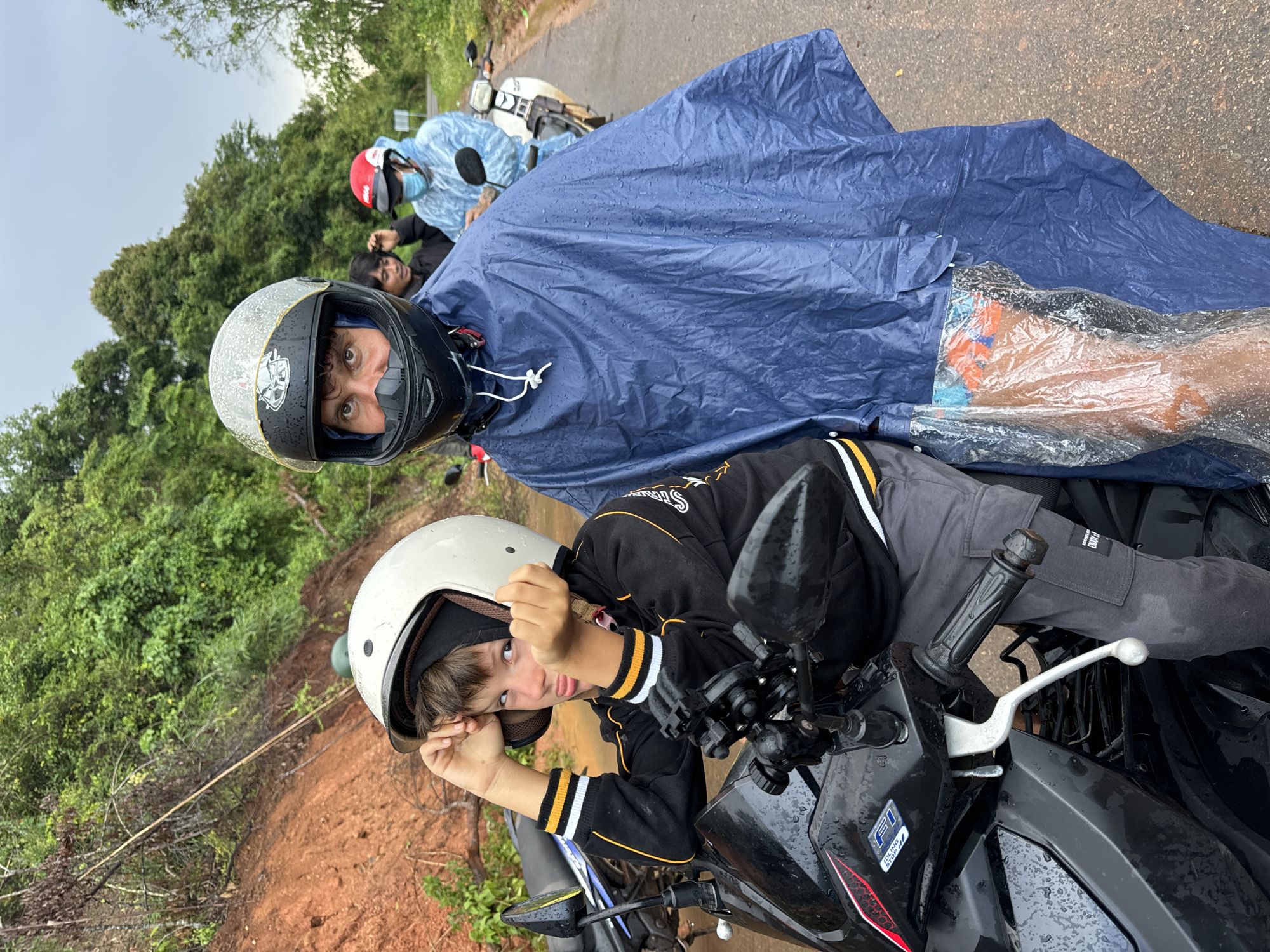 A group of people are riding motorbikes on a wet, paved road in a rural, forested area. The main subjects are two individuals in the foreground. A young boy wearing a white helmet and a black and yellow jacket is on the left, leaning forward on his motorbike and looking at the camera with a playful expression. To his right, a man in a black helmet and a blue poncho is also on a motorbike, looking directly at the camera. The man's poncho is covering most of his body and the motorbike he is on. In the background, other people on motorbikes are visible, including one wearing a red helmet and a light blue rain jacket, and another person with dark hair wearing a black jacket. The environment is lush with green trees on either side of the road, and a reddish-brown landslide is visible on the left side of the road, suggesting it might be the side of a hill or mountain. The road surface is wet and reflective, indicating recent rain. The sky is overcast, contributing to the muted lighting of the scene. The image appears to have been taken in Vietnam, given the context of the motorbikes and the rural landscape.