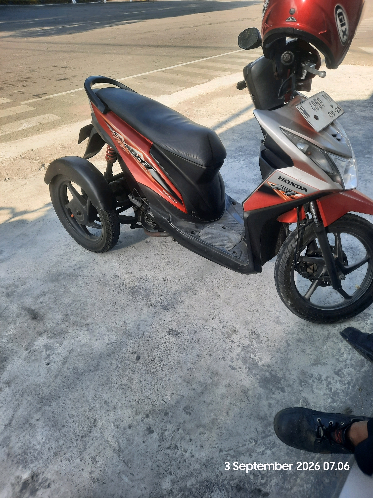 A modified red and silver Honda Beat scooter, with a third wheel attached to the rear, is parked on a concrete surface next to a paved road. The scooter has a red helmet resting on its handlebars. The license plate reads "N 4559 ND 08-28". In the foreground, a person's legs and black shoes are visible. The scene appears to be in Gending, Indonesia, as indicated by the timestamp "3 September 2026 07.06". The environment is outdoors, with shadows cast by the scooter and helmet on the concrete.