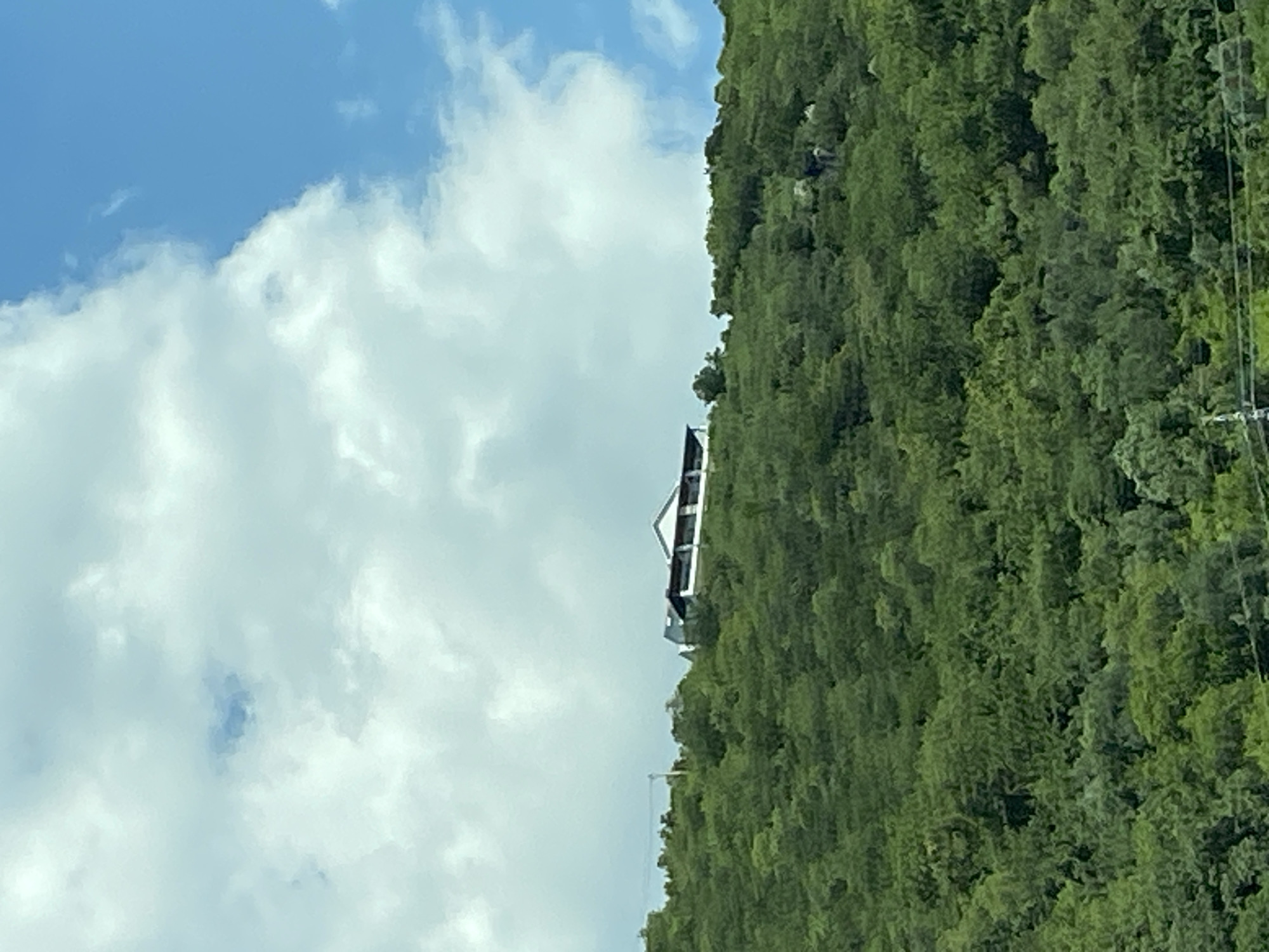 A house with a white facade and a dark roof sits on a lush, green hillside under a blue sky with scattered white clouds. The house appears to be a single-story structure with a covered patio or balcony along the front. The hillside is densely covered with trees, creating a verdant landscape. The scene captures a serene view of a home nestled in nature.