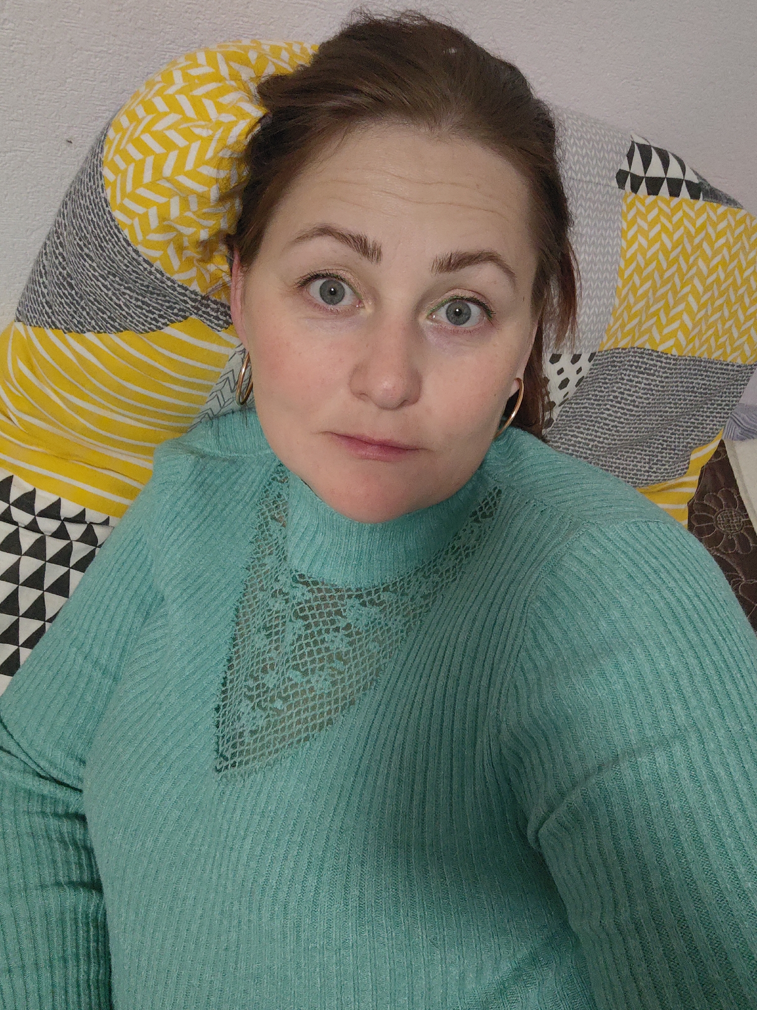 A close-up portrait features a woman reclining indoors, looking directly into the camera. Her head and upper torso are visible, positioned against a patterned pillow or cushion and a plain light-colored wall.
The woman has light reddish-brown hair pulled back from her face. Her eyes are wide, light in color (appearing blue-grey or greenish-blue), and focused, giving her a contemplative or slightly intense expression. Faint lines are visible on her forehead, and her eyebrows are well-defined. She has a subtle pink tint on her lips and cheeks and is wearing gold-colored hoop earrings.
She is dressed in a long-sleeved, ribbed sweater in a vibrant light turquoise or teal color. The sweater features a distinct open-knit or lace pattern in a V-shape across the upper chest, under a mock neckline, adding a decorative element to her attire.
Behind her head, a pillow or cushion displays a bold geometric pattern composed of various sections: bright yellow with white chevron stripes, black and white triangles, and grey areas with textured lines. The overall impression is a casual, intimate moment captured in a home setting. The lighting is even and bright, suggesting an indoor daytime photo or good artificial illumination. There is no visible text in the image.