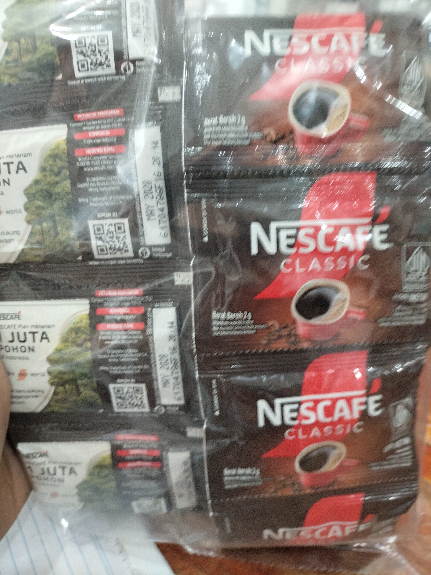 The image displays several individual sachets of Nescafé Classic coffee, bundled together. The sachets are primarily black and red, featuring the Nescafé logo in white lettering. Each sachet also has an image of a red coffee cup filled with coffee and coffee beans. Some text is visible on the sachets, including "NESCAFÉ CLASSIC," "Berat Bersih 2g" (Net Weight 2g), and expiration dates like "MAY 2026." There are also QR codes and what appear to be nutritional or ingredient information in Indonesian on some of the sachets. The sachets are wrapped in clear plastic. One sachet on the left side of the image also features a campaign related to planting trees, with text like "JUTA POHON" (Million Trees) and "our world." A hand is visible at the bottom left of the image, partially obscuring the sachets. The background is out of focus but appears to be indoors. There is no discernible information in the image that places it specifically in the city of Meudangara, Indonesia.