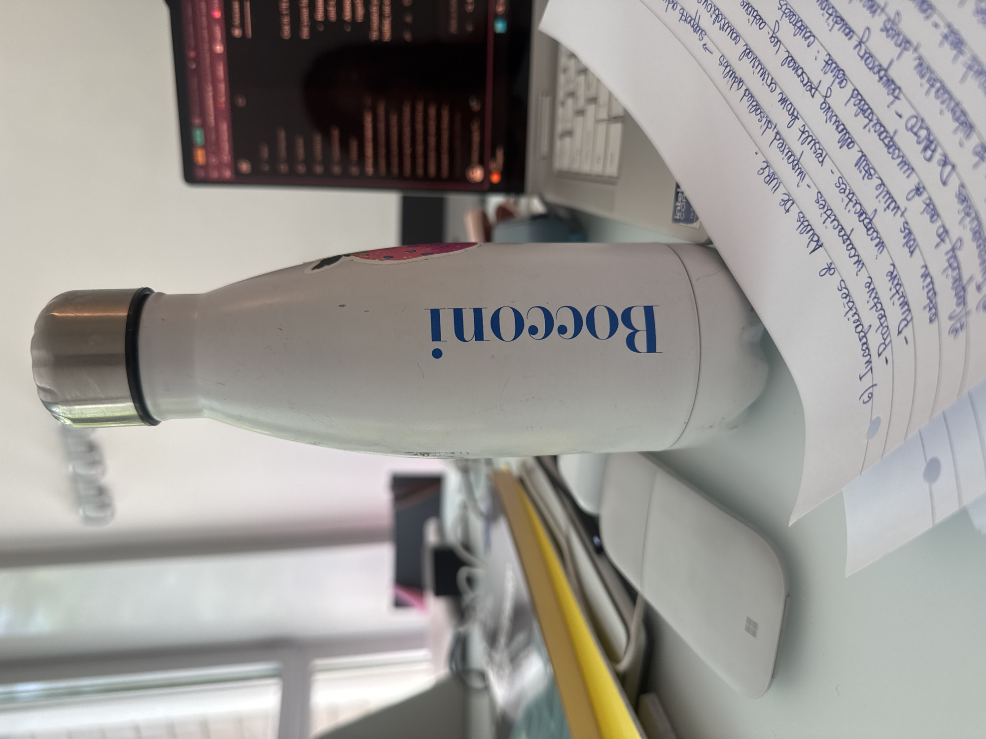 The image shows a white "Bocconi" water bottle lying on a desk. The word "Bocconi" is printed in blue on the bottle. The water bottle is next to a white Microsoft mouse and a stack of papers with handwritten notes in blue ink, possibly Italian due to the presence of the term "incapacities DE IURE". The notes seem to be about legal or academic topics. In the background, there's a blurred view of a computer screen displaying data, and part of a window. Given the presence of the "Bocconi" water bottle, it's likely this scene is located in the city of Milan, Italy, near Bocconi University.
