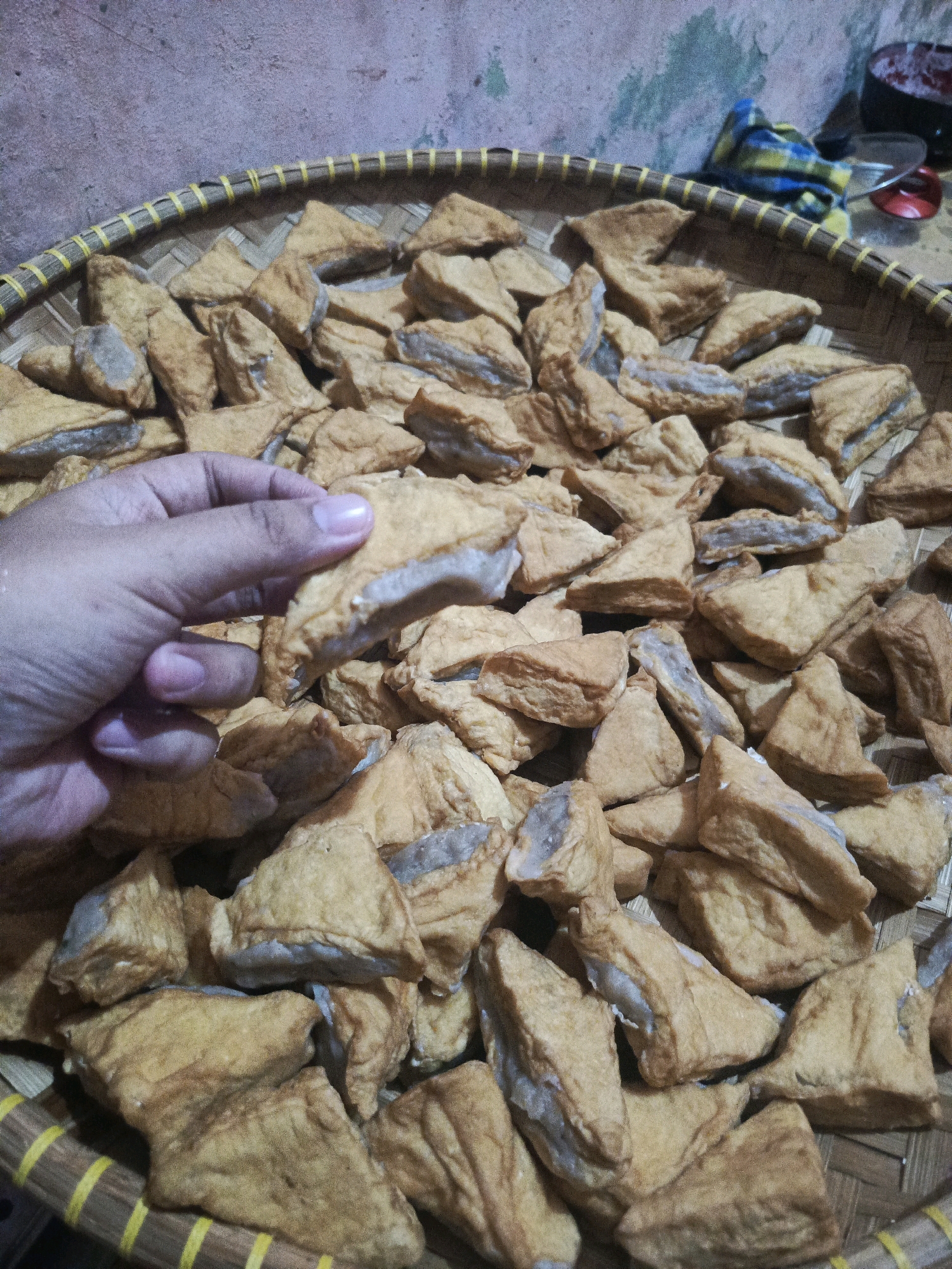 A close-up, top-down shot shows a hand holding a triangle-shaped piece of fried tofu over a woven bamboo tray filled with more fried tofu. The tofu pieces are golden brown and appear to be seasoned. The background is a textured, light-colored wall. The scene is likely set in Balaraja, Indonesia, a region known for its agricultural products and local cuisine.