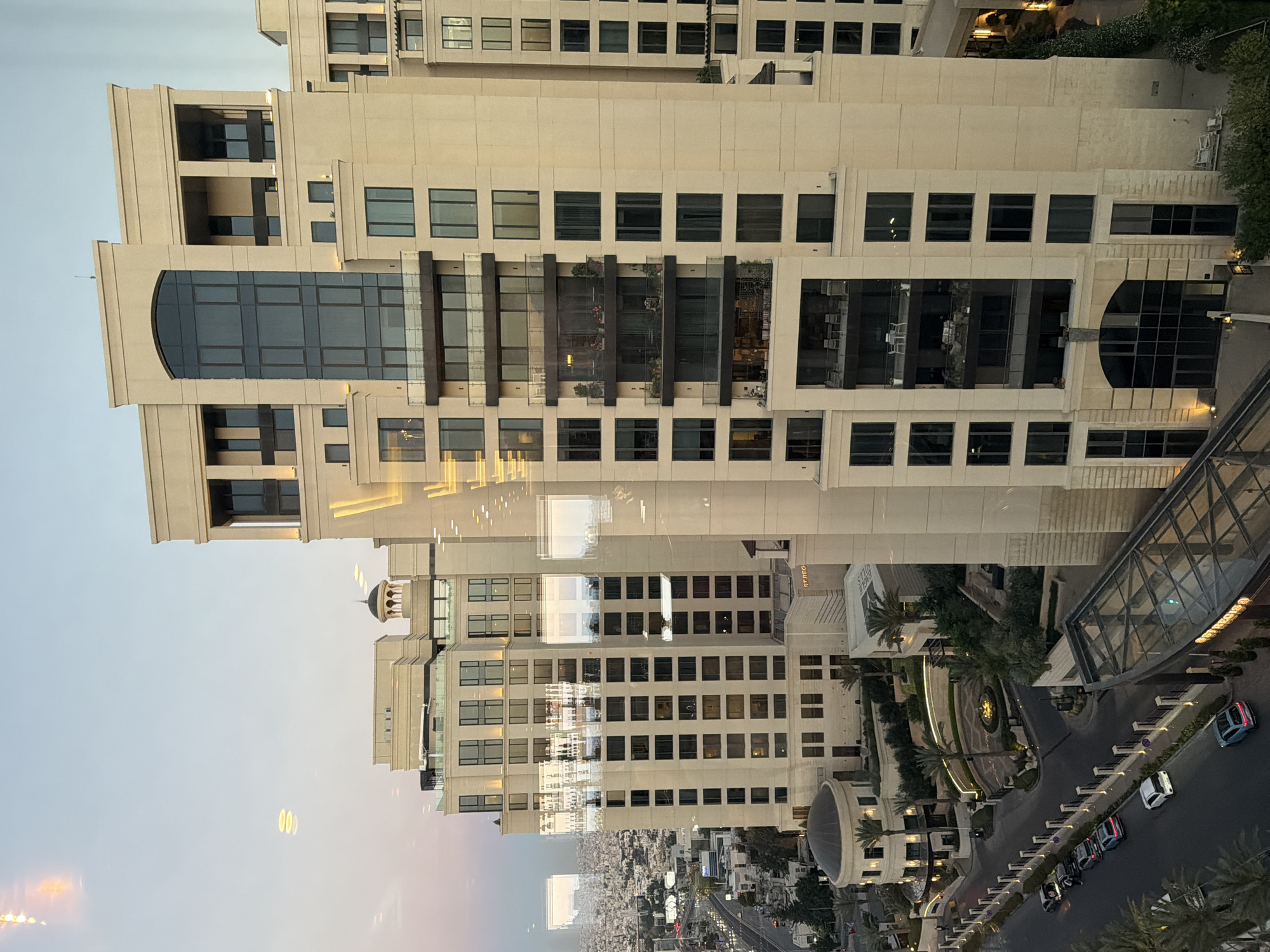 The image shows a large, modern hotel complex in Amman, Jordan, likely during the late afternoon or early evening. The primary focus is on the architecture of the hotel, characterized by its cream-colored facade, numerous windows, and several balconies. The hotel appears to be composed of multiple connected buildings of varying heights, with a taller, central tower.
In the foreground, a glass-covered walkway leads to one of the hotel entrances. Below the hotel, a street is visible with several cars parked along the side. The street is lined with decorative fencing and landscaping, including palm trees and manicured bushes. Further in the distance, the cityscape of Amman stretches out, with a dense collection of buildings under a pale sky. Reflections are visible on the hotel windows, suggesting the photograph was taken from another building or through a window. The lighting in the scene is soft, indicative of twilight. There are no clearly identifiable people or animals in the frame.