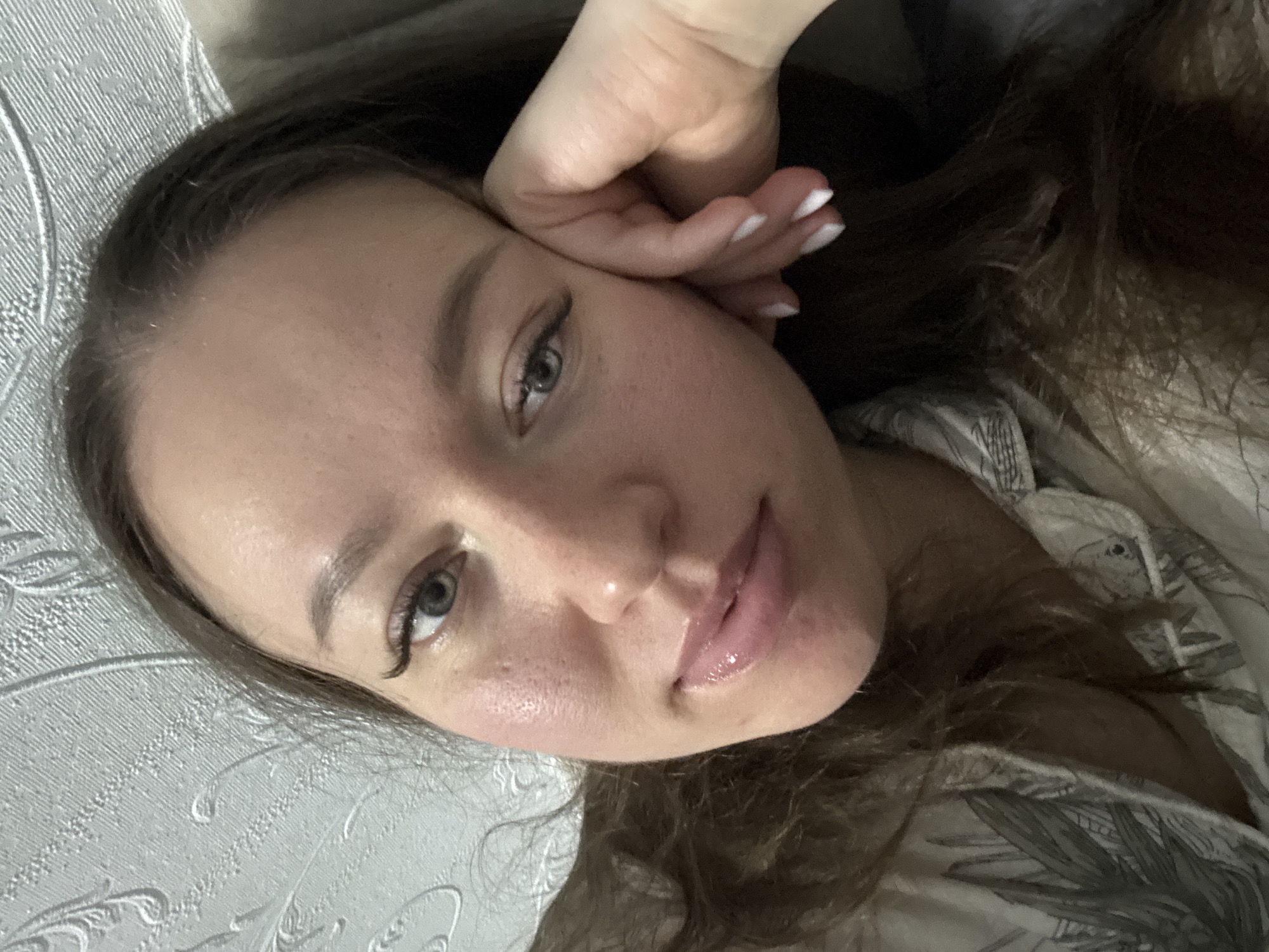 This media file is a close-up portrait of a young woman, likely a selfie, taken indoors. She is lying on her side, looking directly at the camera with her head slightly tilted. Her right hand, with fingers curled and nails painted white, supports her head near her temple and cheek.
The woman has fair skin, long dark brown hair, and light-colored eyes, appearing grey-green. She wears winged eyeliner and mascara, and her full lips are glossed with a natural pink hue, slightly parted. Her expression is calm and relaxed, with a soft, gentle gaze.
Her head rests on a light gray surface, which appears to be a pillow or bedding, featuring a textured, embossed pattern of swirls and floral designs. On the right side of the frame, partially obscured by her hair, is a light-colored fabric with a distinct pattern of dark green or grey leafy motifs, possibly part of a shirt or duvet cover.
The lighting is soft and even, consistent with an indoor setting. No specific activity beyond posing for the photograph is depicted. There is no visible text in the image. Although the location context "Ферма, Russia" was provided, there are no explicit visual cues within the image itself to confirm this specific geographical location; the scene suggests a domestic interior.
