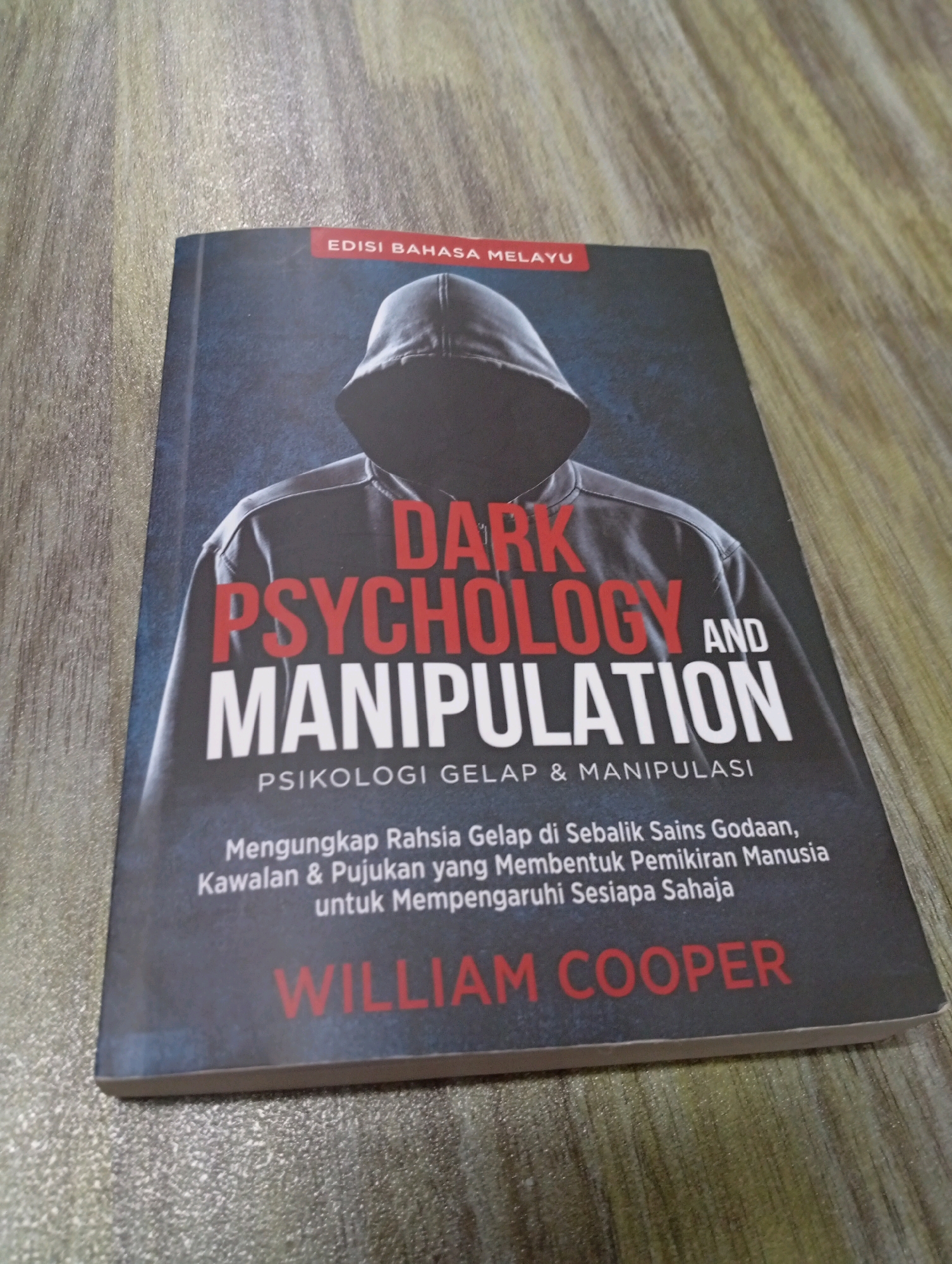 A book with a hooded figure on the cover is lying on a wooden surface. The book's title is "DARK PSYCHOLOGY AND MANIPULATION" in large red and white lettering. Below the title, in smaller white text, is "PSIKOLOGI GELAP & MANIPULASI". Further down, a description in Malay reads: "Mengungkap Rahsia Gelap di Sebalik Sains Godaan, Kawalan & Pujukan yang Membentuk Pemikiran Manusia untuk Mempengaruhi Sesiapa Sahaja". At the bottom of the book, the author's name, "WILLIAM COOPER", is printed in red. A red band at the top of the cover says "EDISI BAHASA MELAYU". The image is a close-up of the book, with the wooden surface providing the background. There are no people visible in the image, and the setting appears to be indoors, likely a desk or table. The book is the primary subject. There is no information to suggest the image was taken in the city of Selayang Municipal Council, Malaysia.