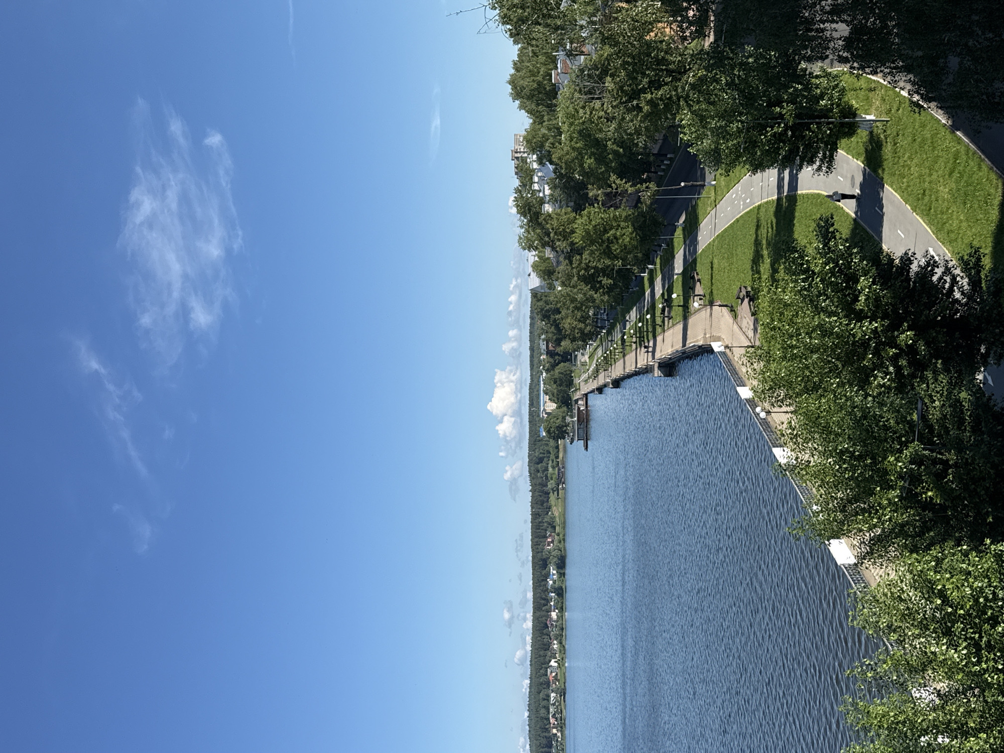 The image captures a vibrant, sunny daytime scene overlooking a wide body of water, likely a reservoir or large lake. The sky above is a brilliant, clear blue with a few wispy, scattered white clouds.
In the foreground, a well-maintained embankment runs along the right side of the water. This bank features a paved path, appearing to be a bike or walking trail, marked with white lines. Lush green grass borders the path, which is also lined with numerous green trees and regularly spaced streetlights. One person is visible walking away from the viewer on this path. Structures resembling small docks or jetties extend into the water from this near bank.
The expansive body of water, a deep blue, shows gentle ripples across its surface, reflecting the bright sky.
On the distant left bank, a long, tree-lined shoreline is visible, behind which several houses and buildings are nestled. The distant horizon also shows faint signs of more clouds.
The scene conveys a peaceful and pleasant outdoor atmosphere, typical of a warm, clear day, with bright sunlight creating distinct shadows from the trees onto the path. No visible text is present in the image. The setting suggests a recreational area along a significant waterway.