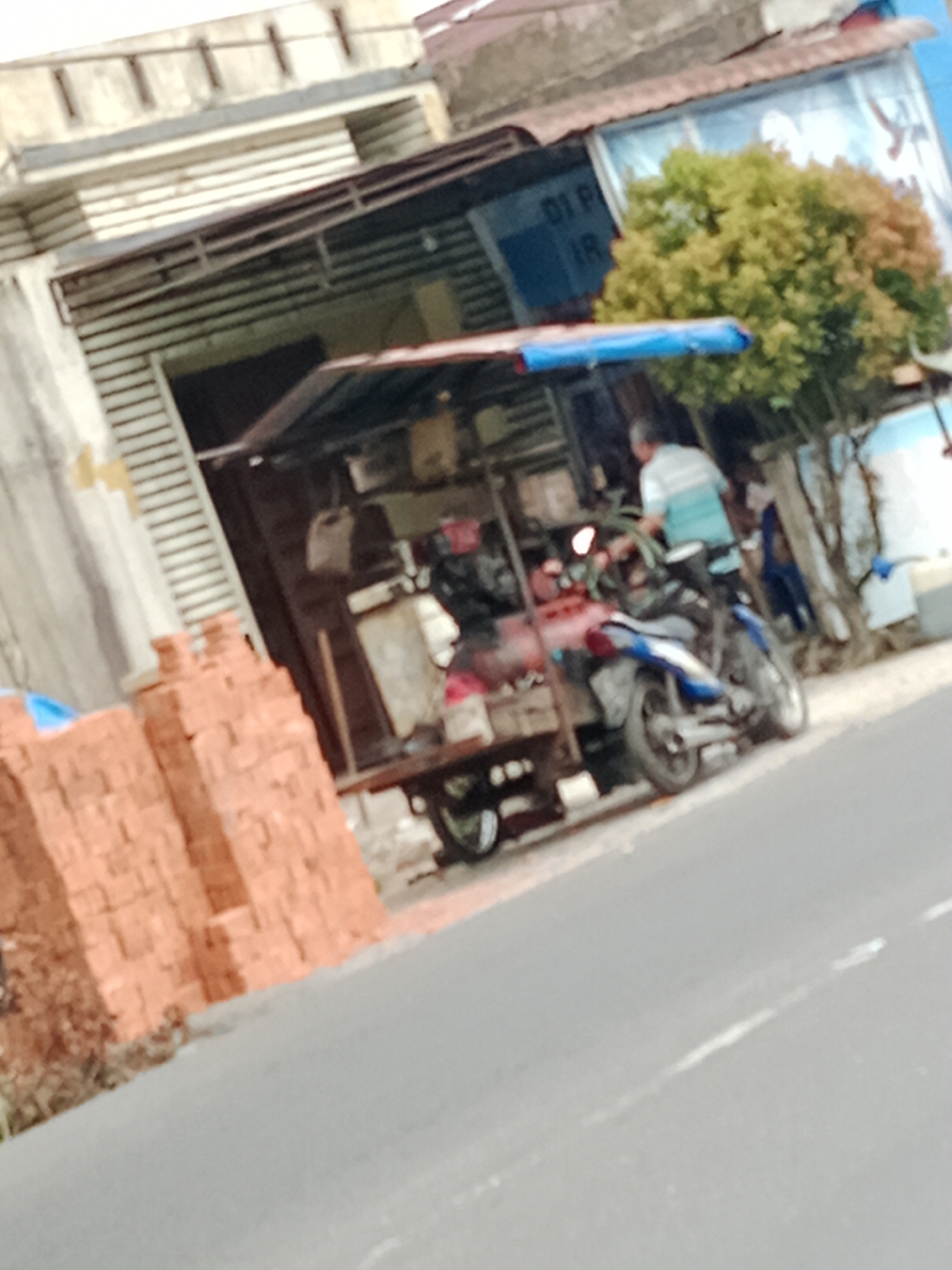 In a street scene in Binjai, Indonesia, a vendor stall is set up, featuring a blue tarp covering part of the structure. In front of the stall, a blue and white motorcycle is parked. A man wearing a striped shirt stands near the motorcycle, possibly interacting with the stall or the vehicle. To the left of the stall, a stack of red bricks is visible, suggesting construction or sale of building materials. The background shows a building with horizontal wooden planks and a tiled roof. A green tree with yellow and green foliage stands behind the stall. The scene appears to be on the side of a paved road, with a white dashed line visible on the road surface.