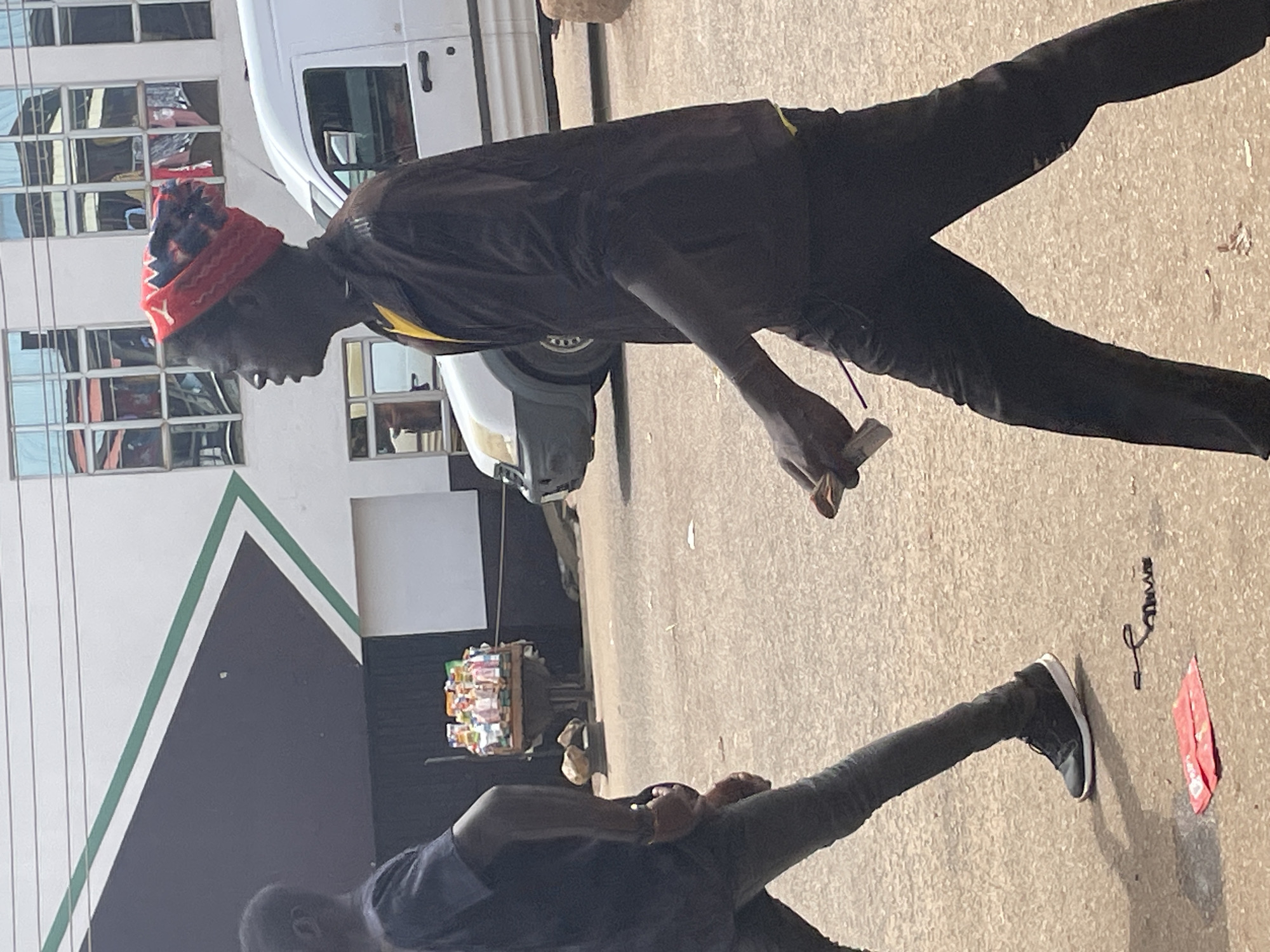 The image shows two young Black men in Bukuru, Nigeria. The man in the foreground is wearing a dark blue shirt and dark pants, and is seated on the ground. The man in the background is standing and wearing a red and black beanie, a dark jacket over a yellow shirt, and dark pants. He is holding a small white object in his right hand, possibly a phone or cigarette.
The scene appears to be outdoors, with a paved ground and a building with reflective windows in the background. There is also a white van parked nearby and a small cart piled with various goods, likely for sale, suggesting a street vendor setup. The lighting suggests it is daytime, likely with bright sunlight casting shadows. The overall atmosphere seems casual, with the two individuals engaged in relaxed activity.
There is no visible text in the image.