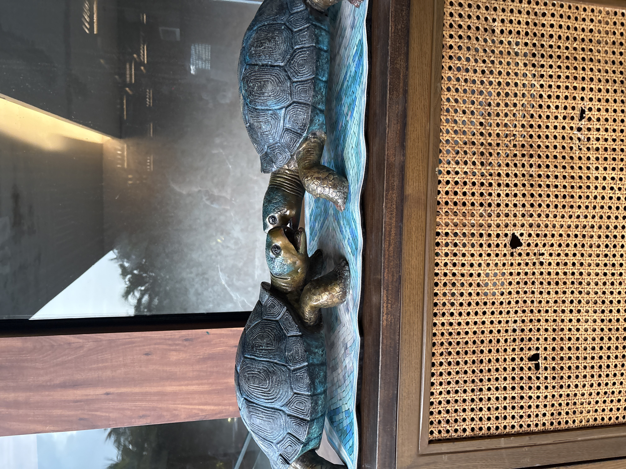 Two bronze turtle sculptures are positioned nose-to-nose on a blue mosaic tile mat placed on a wooden cabinet. The turtles have dark green shells with a bronze-colored head, legs, and undersides. The left turtle's mouth is slightly open, while the right turtle's mouth is also open, appearing to be kissing or interacting with the other. The cabinet has a dark wood finish and a woven cane front with some minor damage. Behind the cabinet, a large window reflects the sky and palm trees, suggesting an outdoor or semi-outdoor setting. The overall scene appears to be a decorative display. This is likely located in Vagator, India, given the context.