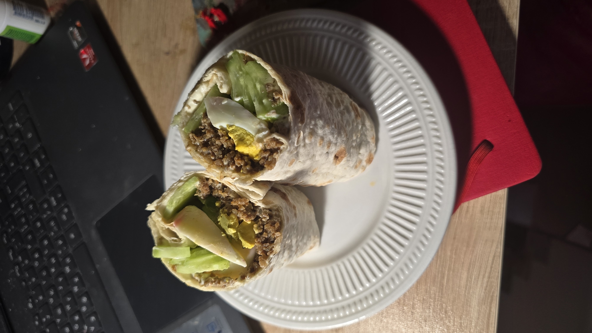 On a wooden table sits a white plate holding a sliced burrito, next to a black laptop. The burrito appears to contain ground meat, cucumber slices, egg, and some kind of sauce. The plate is partially resting on a red notepad. There's also a small, green and white bottle visible in the upper left corner near the laptop. The location is likely indoors, but there are no distinct features that suggest the city of Stara Zagora, Bulgaria.