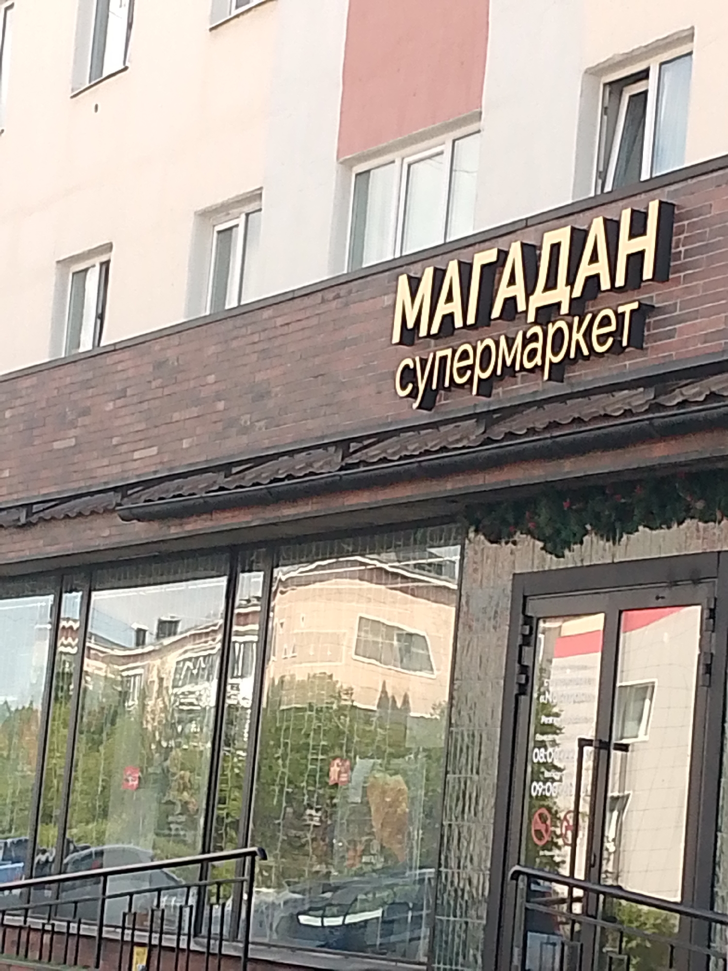 The image shows the facade of a supermarket named "МАГАДАН" (Magadan). The name is displayed in large, golden, three-dimensional letters with black outlines on a brick wall above the entrance. Below the main name, smaller, golden letters spell out "супермаркет" (supermarket). The building has multiple windows, some of which are open. Reflections of other buildings and trees are visible in the large glass windows of the supermarket. There are also signs on the glass doors indicating opening hours. The scene depicts a typical street view of a commercial establishment in a city, likely in Magadan, Russia, given the name of the supermarket.