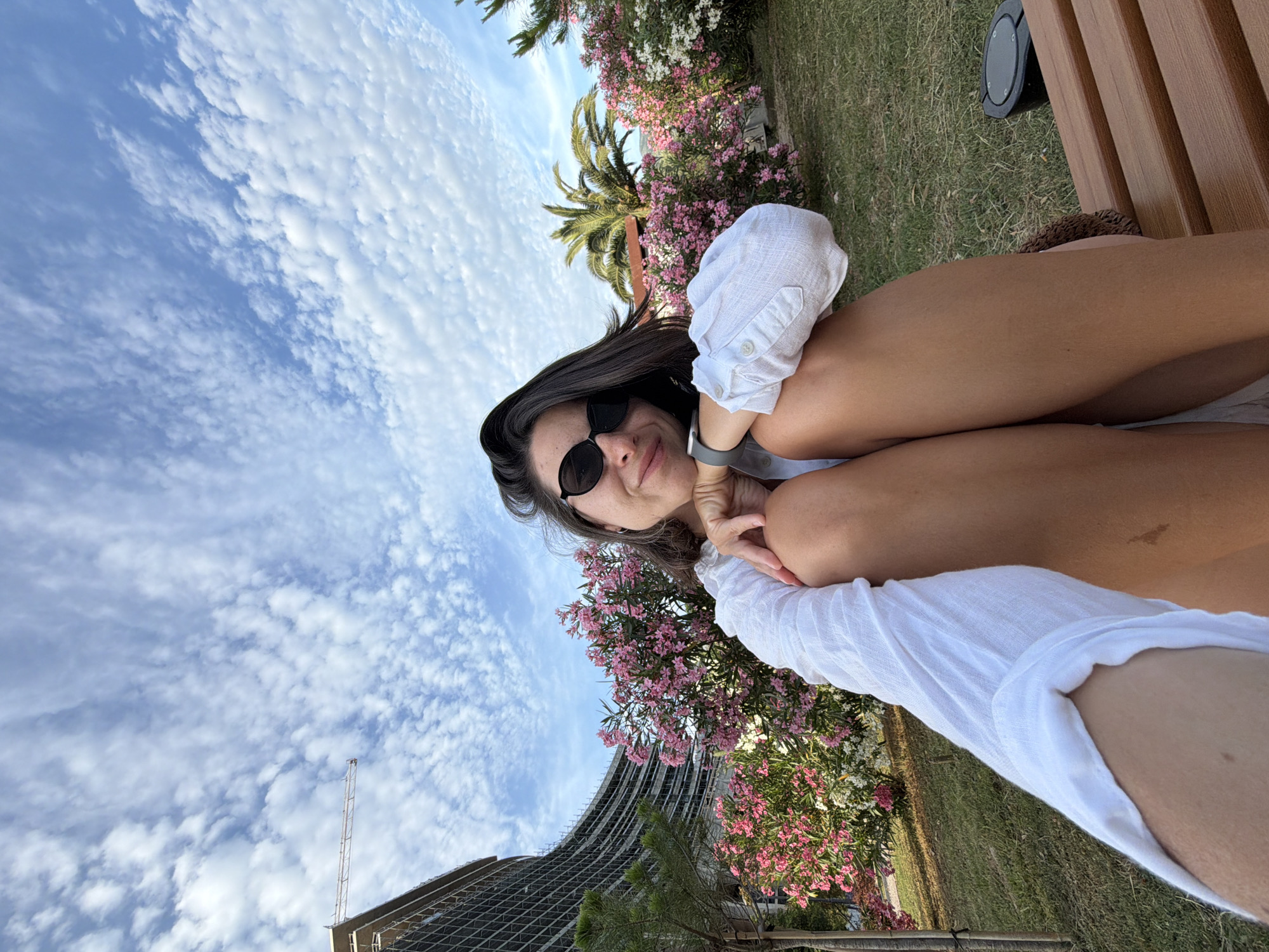 An outdoor, wide-angle selfie captures a young woman sitting on a brown wooden slatted bench under a bright blue sky with scattered cumulus clouds in Budva, Montenegro. The woman, with dark hair and wearing black sunglasses and a white long-sleeved shirt, smiles directly at the camera. Her arms are bent, resting on her knees. A dark smartwatch is visible on her left wrist. To her left, a large, multi-story, curved building under construction is prominent, with scaffolding covering its exterior, and a construction crane extends from its upper left side. In the mid-ground, vibrant pink and white oleander shrubs are in bloom, interspersed with green foliage and palm trees. Green grass covers the ground, and a small, dark circular ground light fixture is visible to the right of the woman's legs. The scene is illuminated by natural daylight.