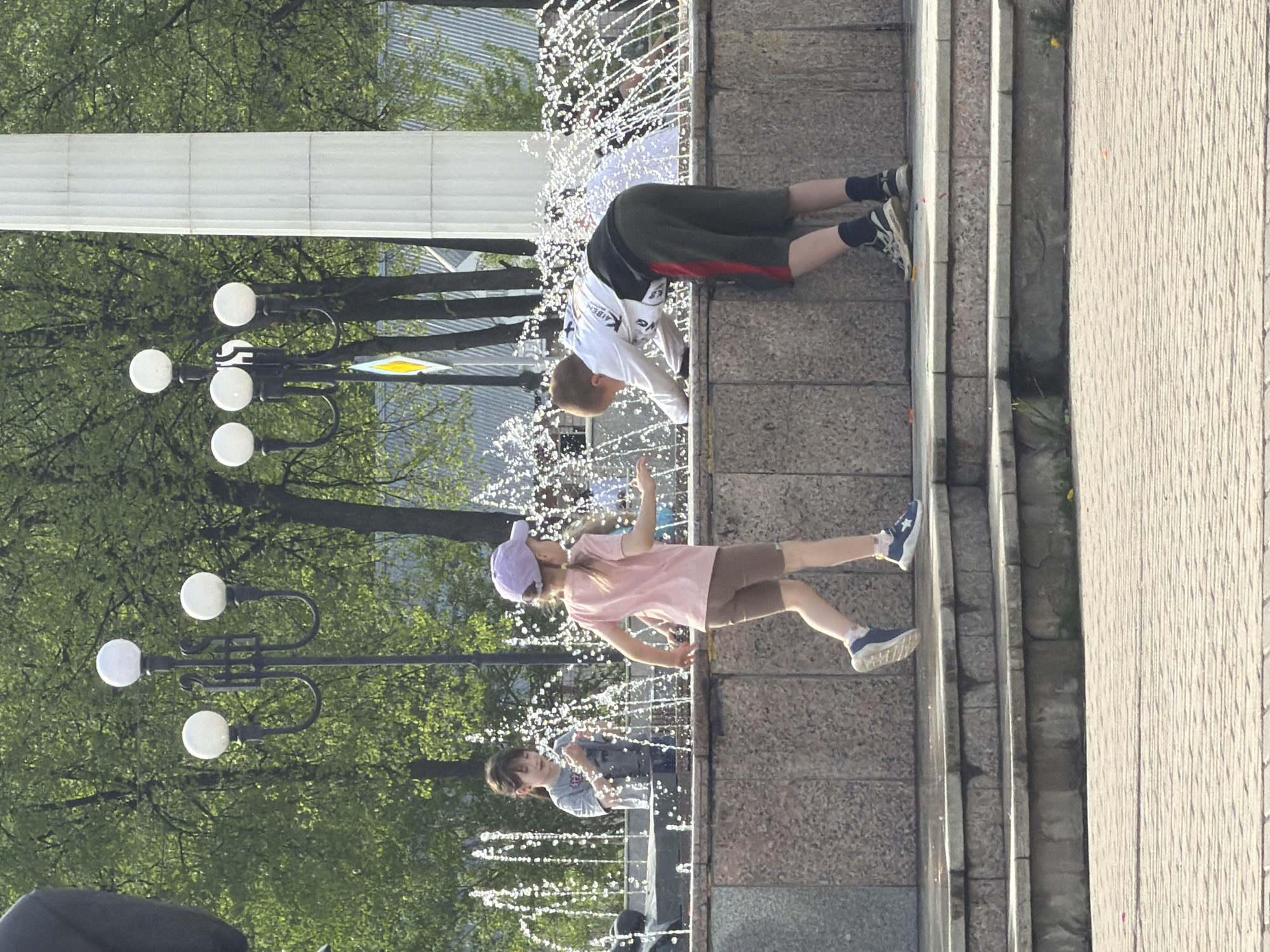 The image shows several children playing in a fountain. One child is lying prone with their head under the spray, another is walking through the water, and another is visible in the background. The fountain appears to be located on a paved area, possibly a plaza or park, with lamp posts nearby. The image was taken in Chaikovsky, Russia.