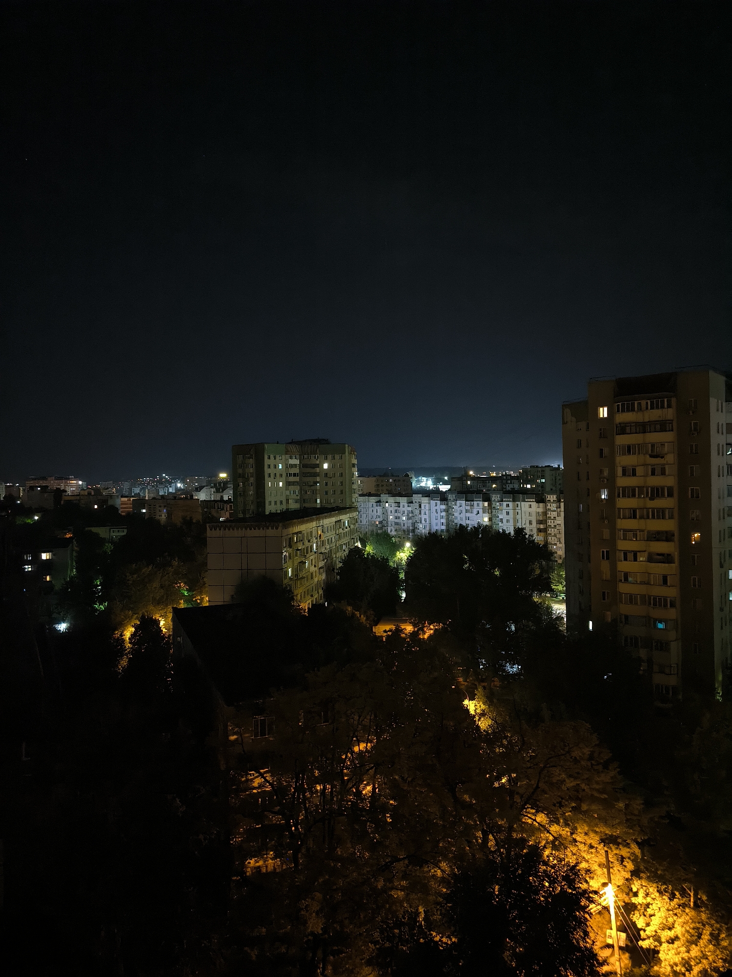 The image is a nighttime aerial view of a residential area in the city of Chișinău, Moldova. The scene is dominated by apartment buildings and trees. The sky is dark, with a few faint stars visible. The apartment buildings, typical of Soviet-era architecture, are illuminated by interior lights, casting a warm glow from their windows. Some buildings are taller and more modern, while others are shorter and appear older. The trees in the foreground are dark silhouettes against the illuminated buildings and the night sky. Yellow streetlights illuminate patches of the foliage and the ground, creating an atmospheric and somewhat eerie effect. The overall impression is one of a quiet, urban night, with the city lights providing the only significant illumination.