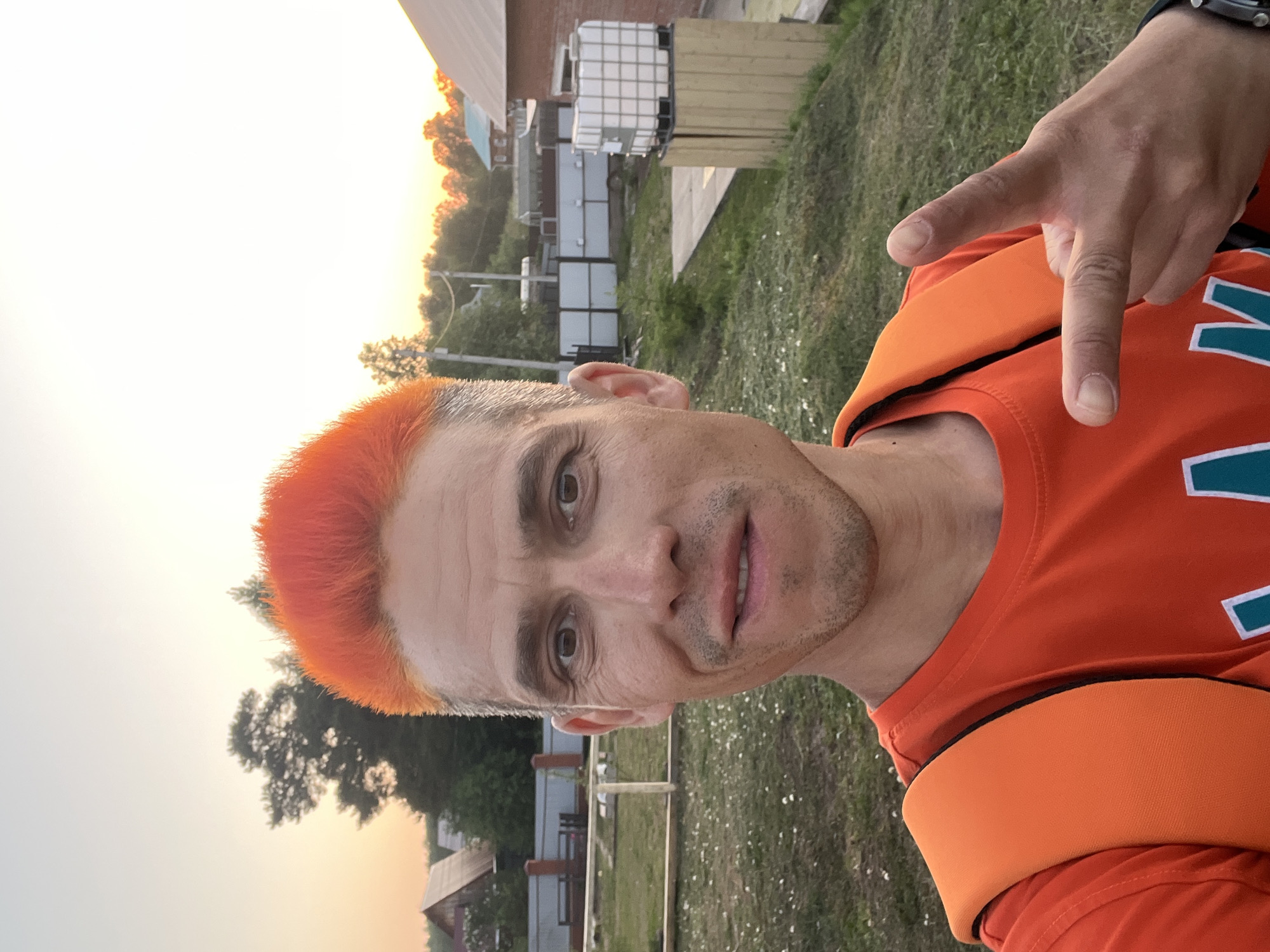 A man with orange hair is making a hand gesture in an outdoor location. He is wearing an orange shirt with teal and white accents and a watch on his wrist. A backpack with orange straps is visible. The background includes a building, trees, grass, and a sunset. The specific location in the city of Chaikovsky, Russia, cannot be determined from the image.