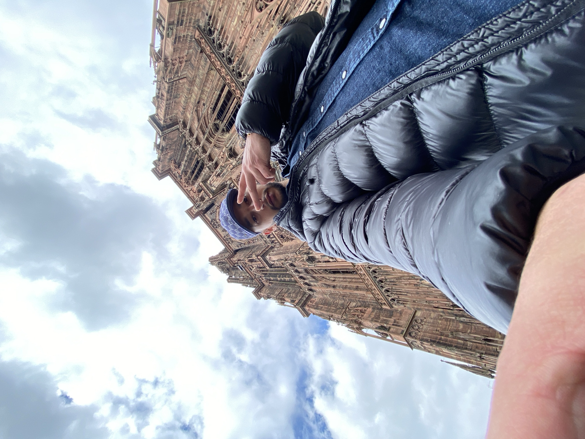 A low-angle, eye-level perspective shows a person in the foreground, wearing a black puffer jacket over a denim shirt and a blue paisley-patterned bandana. The person is flashing a peace sign. The background features the Strasbourg Cathedral, a large, ornate, reddish-brown stone structure with intricate Gothic architecture. The sky is visible, with a mix of white and gray clouds. The location is Strasbourg, France, as indicated by the presence of the Strasbourg Cathedral.