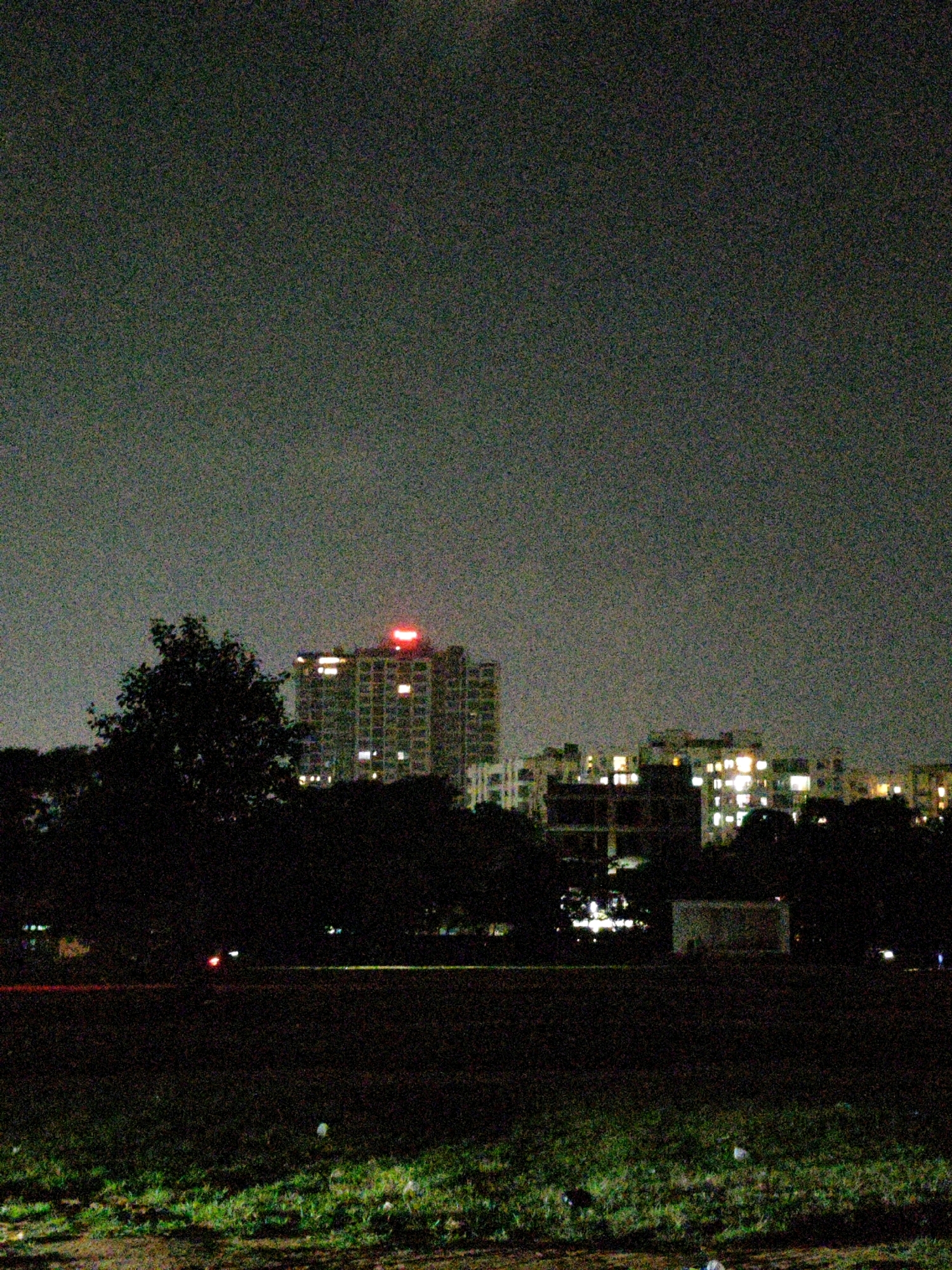 A nighttime exterior shot captures a city skyline in Ranchi, India. The sky is overcast and grainy, with a subtle gradient of light from top to bottom. In the mid-ground, a tall, multi-story building with illuminated windows stands out, topped with a bright red neon sign. To the left of this building, a large, silhouetted tree partially obscures the view. Several other buildings with lit windows are visible to the right, creating a luminous urban panorama. The foreground is dominated by a dark, grassy field. A faint red glow from what appears to be a vehicle's taillights is visible on a dark path running horizontally across the middle of the field. In the far background, a simple, rectangular structure is faintly illuminated, and there are other indistinct lights in the distance. The overall impression is one of urban development at night, with the natural elements of trees and fields contrasting with the built environment.