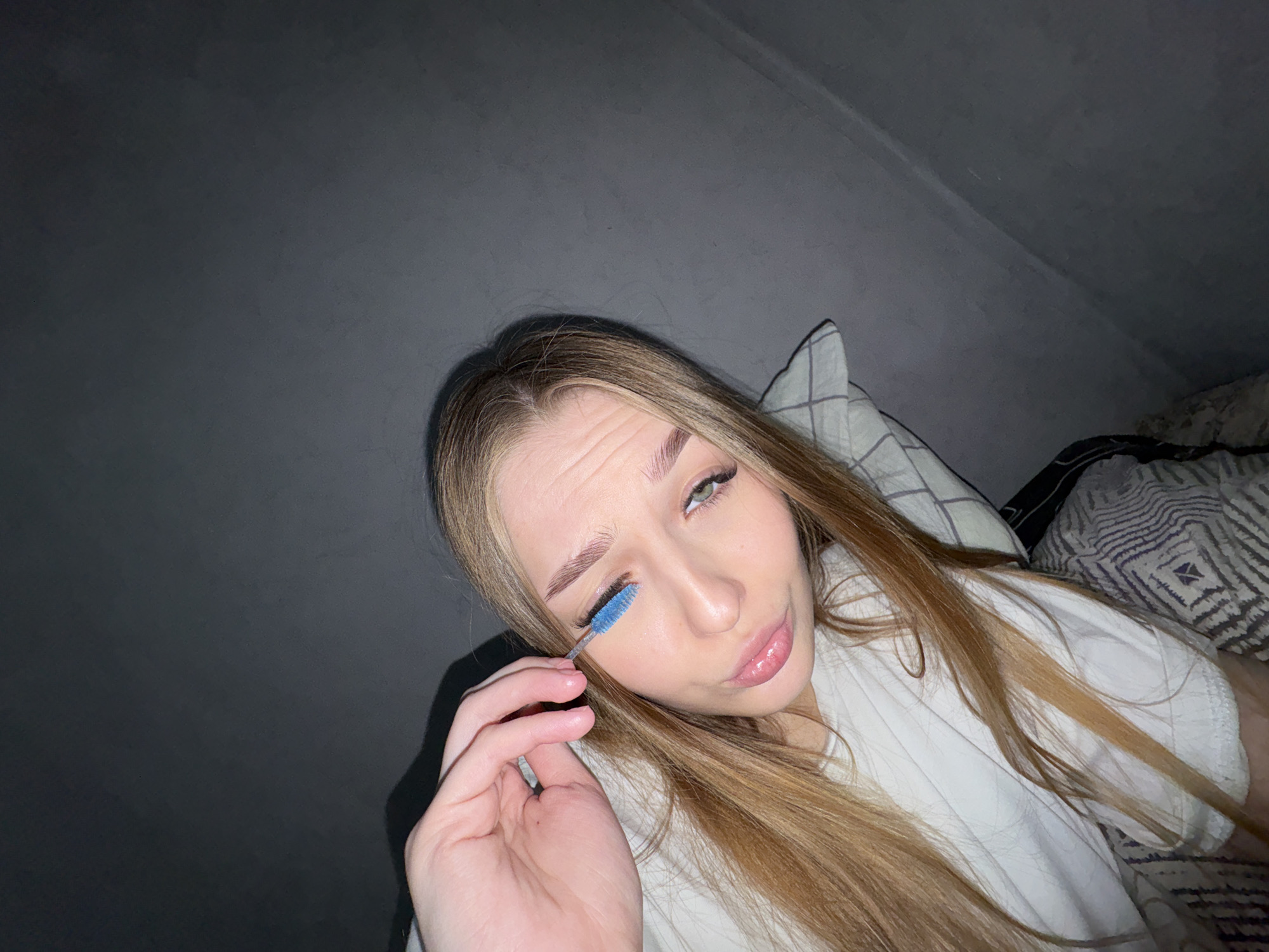 A young woman with long, straight light brown hair is depicted in a close-up, upward-angled shot, likely a selfie. She is lying down or reclining, wearing a simple white t-shirt, against what appears to be a bed with patterned pillows or blankets. Her left hand is visible, holding a bright blue mascara or eyebrow brush, which she is using to brush her right eyebrow. Her left eye is open, revealing a green iris, while her right eye is partially closed as she focuses on the application. She has a slight pout on her lips and her forehead is wrinkled above her right eyebrow, indicating focus or a playful expression directed at the camera. The background is a dark, plain wall or ceiling, possibly angled, suggesting an indoor setting like a bedroom, possibly an attic room due to the sloped wall. The lighting is dim overall, with a direct light source illuminating her face, creating shadows on the wall behind her. No text is visible in the image. This scene takes place in Chaikovsky, Russia.