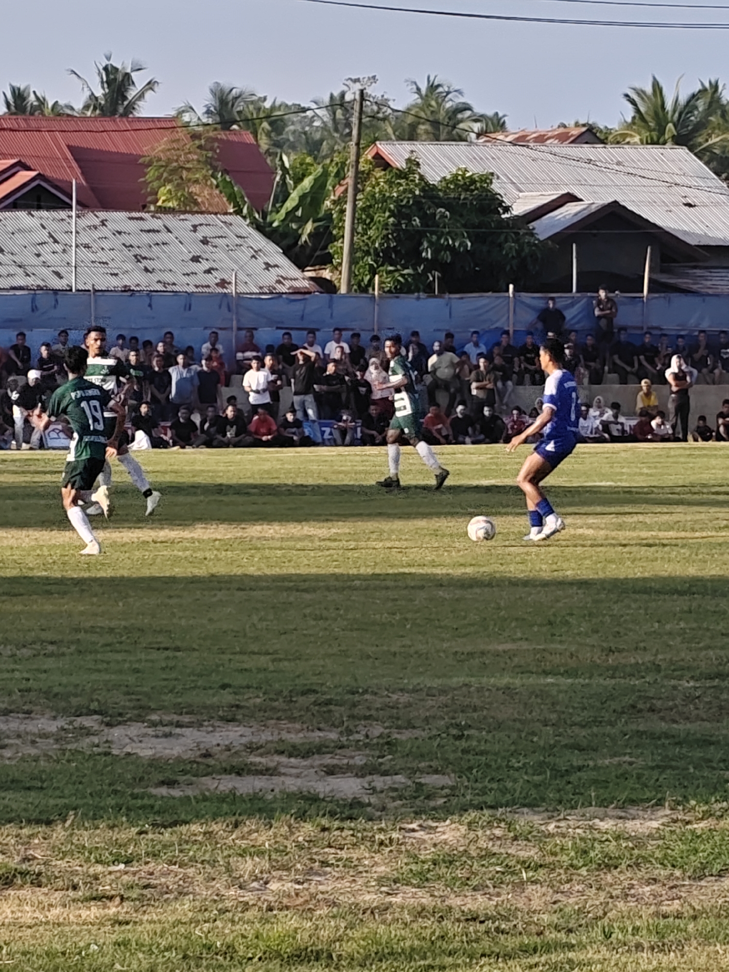 In Desa Meunasah Blang, Indonesia, a soccer match unfolds on a grassy field under a clear sky. A crowd of spectators, dressed in casual attire, are seated and standing behind a blue barrier, observing the game. Two players are in focus on the field: one in a green and white uniform with the number 19, and another in a blue uniform with the number 7. The player in blue is actively engaged with a soccer ball. The background reveals houses with red and corrugated metal roofs, surrounded by lush green trees, indicative of a tropical environment. The overall scene depicts a sporting event taking place in a residential area.
