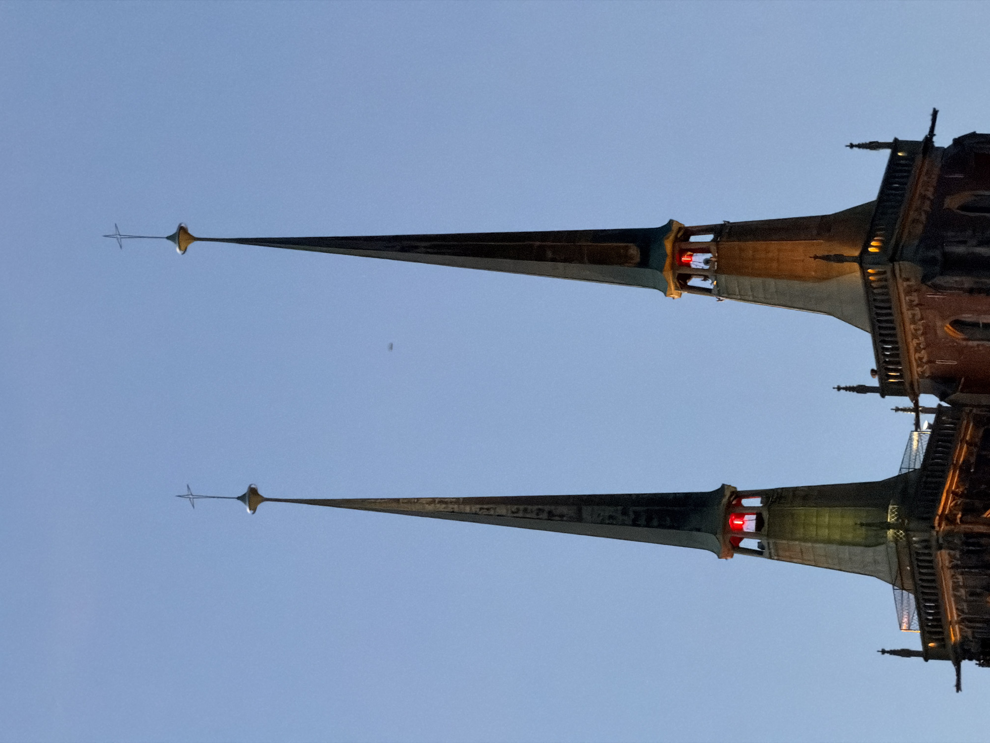 This media file shows a low-angle, outdoor shot of two tall spires, likely part of a gothic-style building such as a cathedral or church. The spires are dark and pointed, with intricate architectural details visible along their bases. Red lights are visible within openings in the structure near the base of each spire, suggesting artificial illumination. Small cross-like ornaments are positioned at the very tips of both spires. The sky is a clear, pale blue, indicating it is either early morning or late evening. There is no text visible in the image. The orientation of the image suggests it was taken at an angle, possibly looking straight up at the spires. The overall mood is serene and awe-inspiring, highlighting the architectural grandeur of the spires against the vastness of the sky.