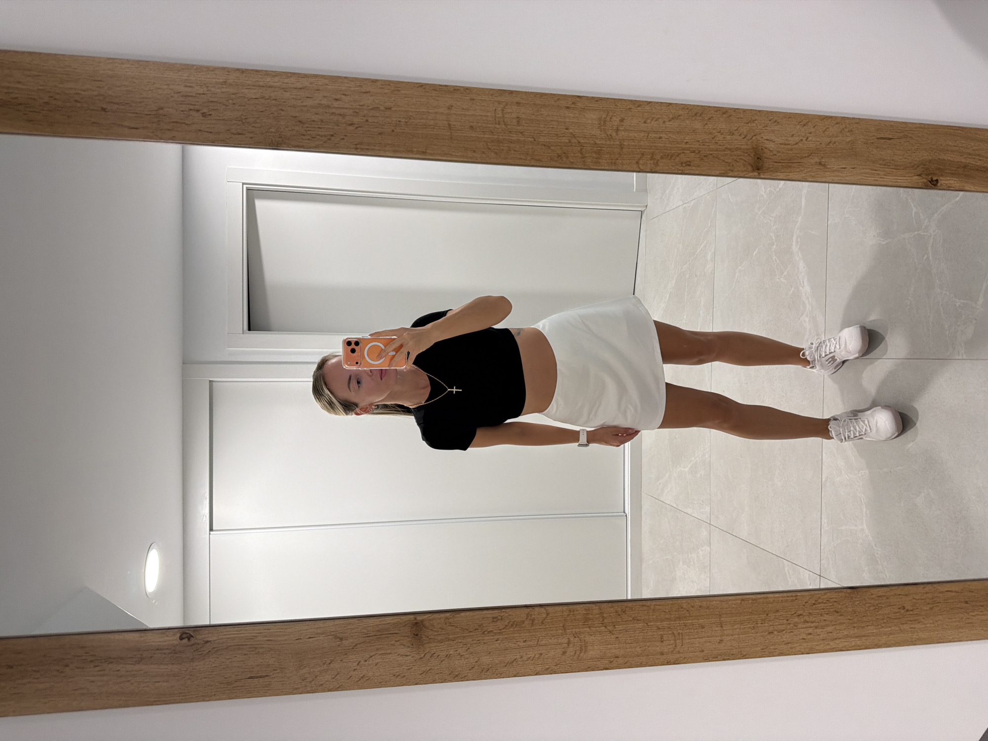 A woman stands in front of a full-length mirror, taking a selfie. She is wearing a black crop top, a white skirt, and white athletic shoes. She has blonde hair pulled back and is wearing a necklace. Her phone, with an orange case, is visible in her hand. The background appears to be an interior space with white walls and a tiled floor. The mirror has a wooden frame. The room seems to be a closet or dressing area. The woman is located in the city of Finestrat, Spain.