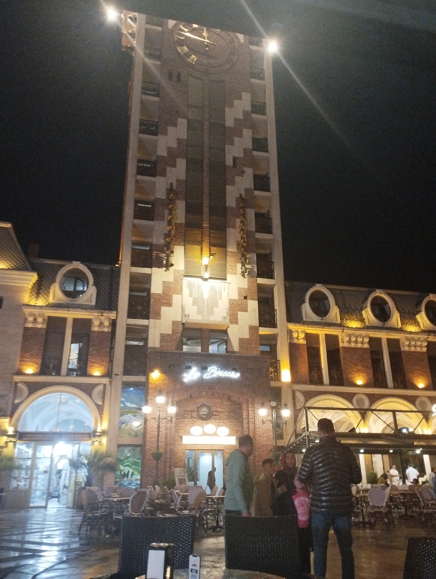 The image depicts a nighttime view of the Piazza Square in Batumi, Georgia. A tall clock tower, decorated with a mosaic pattern, dominates the center of the frame. The tower features a large clock face with Roman numerals. To the right of the tower, an ornate building with arched windows and decorative elements is visible. In front of these structures, an outdoor seating area with tables and chairs suggests a restaurant or cafe. Several people are present in the scene, some standing and conversing, while others appear to be patrons of the establishment. The ground is wet, indicating recent rain, and streetlights illuminate the square. The overall atmosphere is one of a lively urban evening.