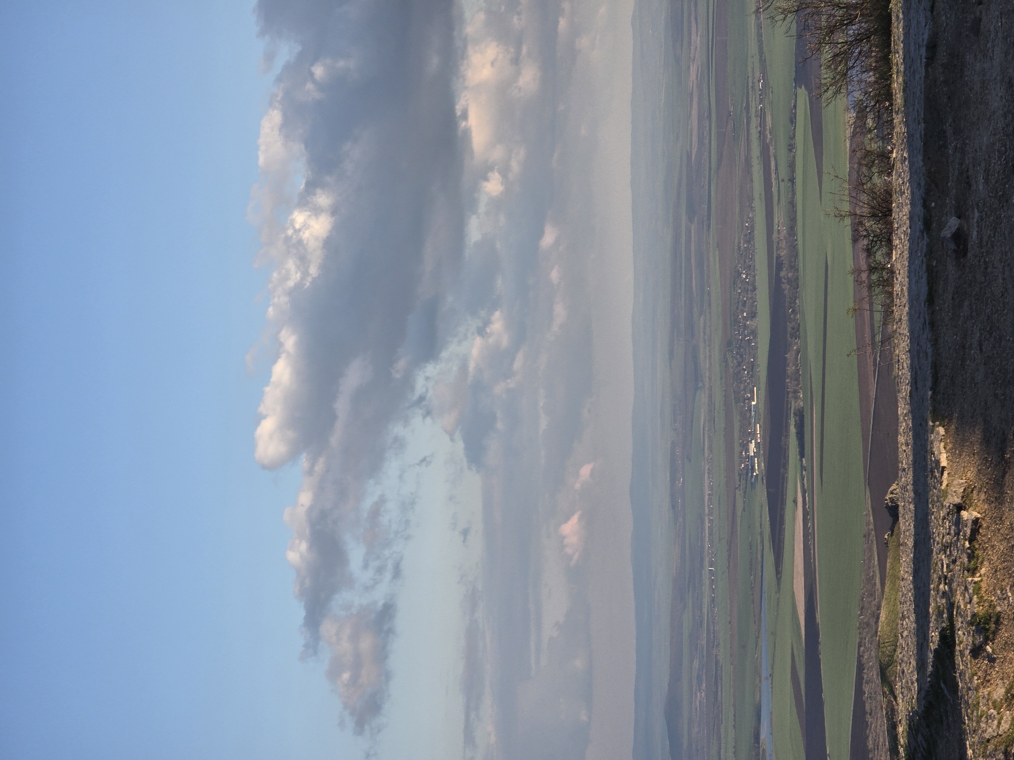The image depicts a rural landscape in Pobeda, Bulgaria, oriented vertically. The sky occupies the left side, transitioning from clear blue to a bank of grey and white clouds. On the right side, a panoramic view of agricultural fields and settlements stretches into the distance. Green and brown patches indicate cultivated land, interspersed with thin lines representing water bodies or irrigation canals. A small settlement is discernible in the mid-ground. A stone wall, with vegetation clinging to its surface, is visible on the right edge, potentially the vantage point of the photograph. Shadows suggest the presence of a setting or rising sun.