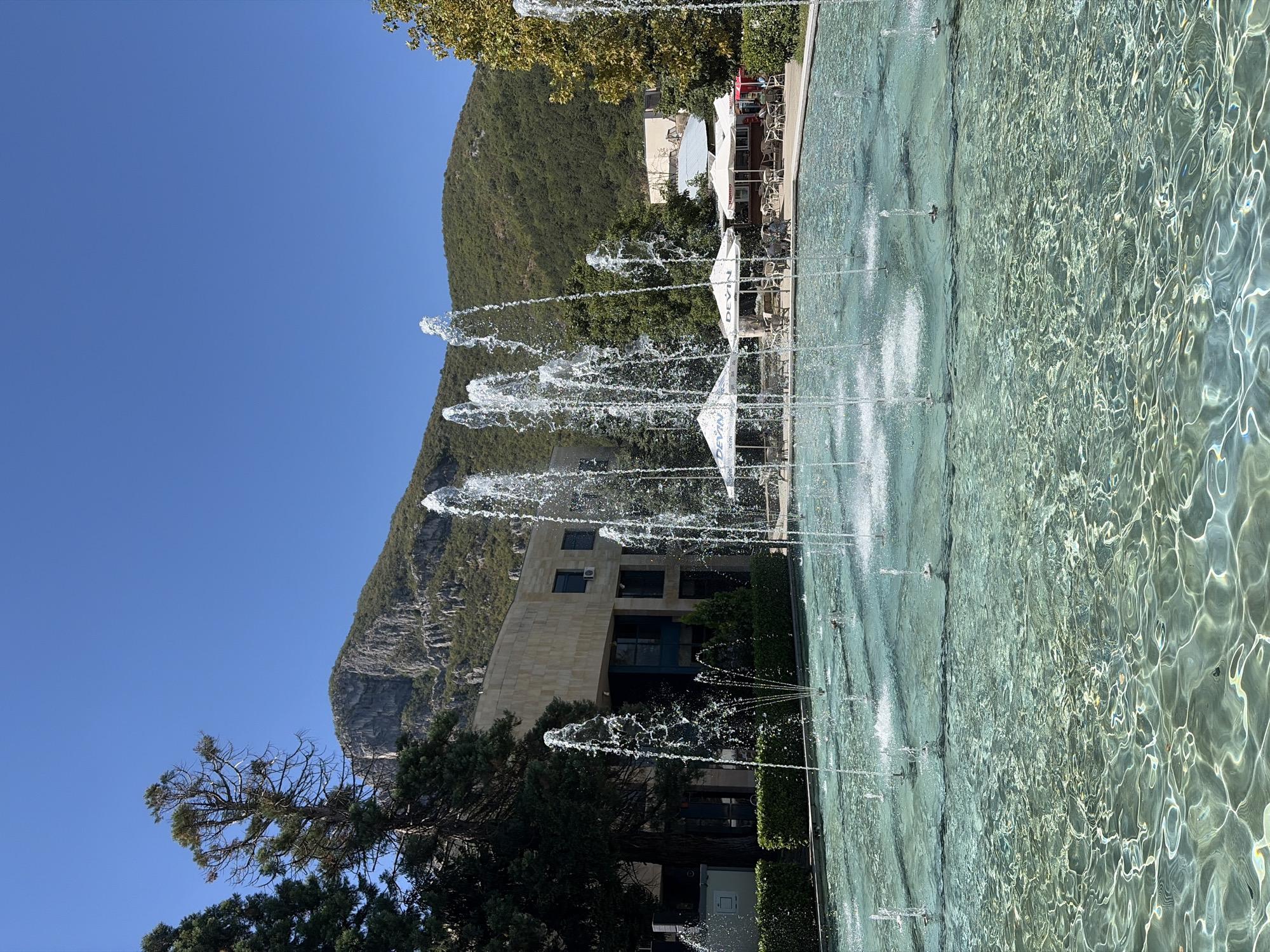 A wide shot captures a public fountain in Vratsa, Bulgaria, on a bright, clear day. The fountain's turquoise water is illuminated by sunlight, creating rippling patterns on the surface. Several streams of water shoot upward from the fountain's basin, some reaching several feet high. In the background, a modern, beige building with dark windows stands behind a neatly trimmed hedge. Beyond the building, lush green trees and a rocky, tree-covered mountain rise under a clear blue sky. To the right, outdoor seating areas with white umbrellas and tables suggest a nearby cafe or restaurant. The scene is peaceful and appears to be a well-maintained public space.