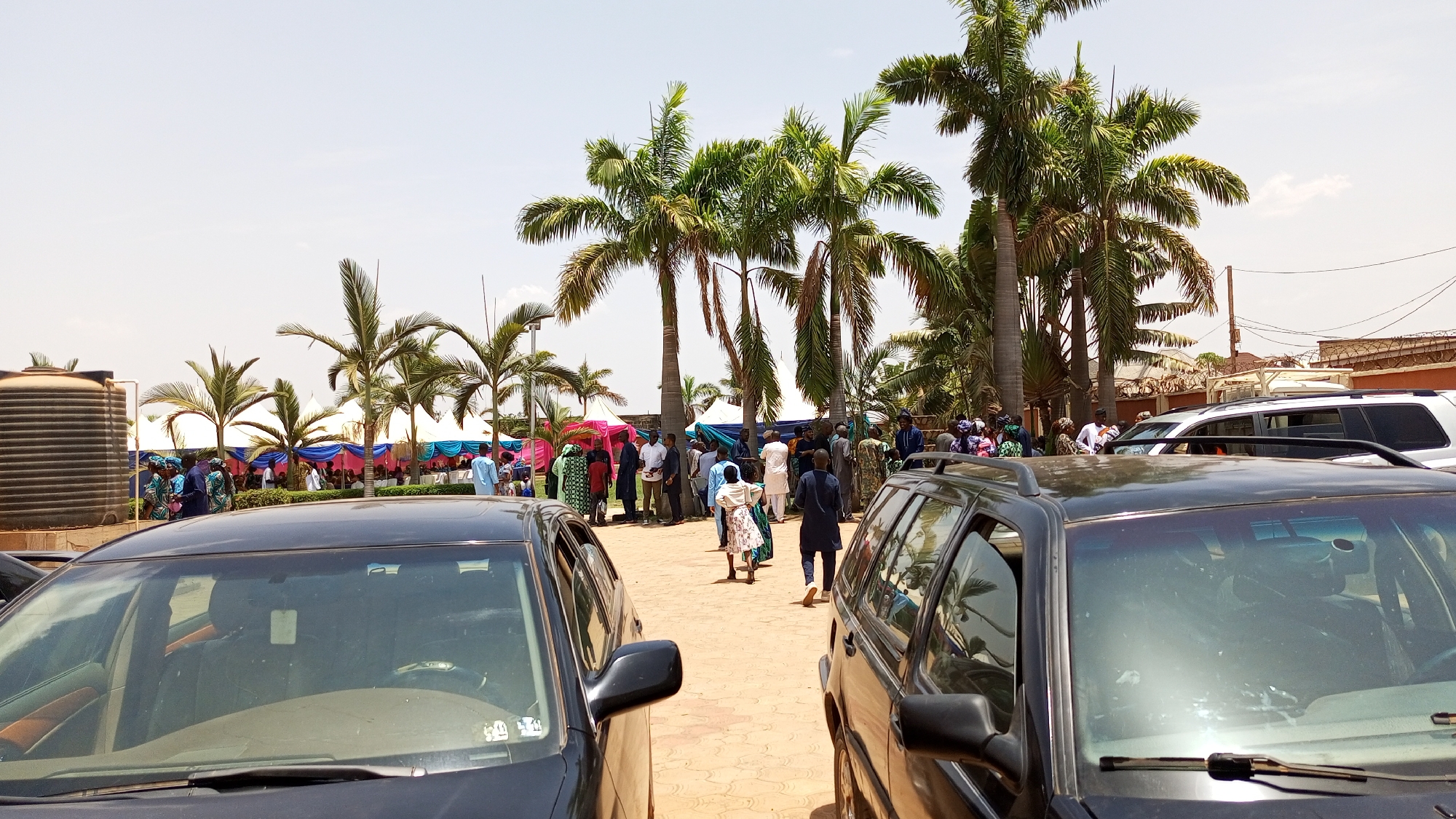 The image, taken during daylight hours in Kakau, Nigeria, shows an outdoor event possibly a wedding or a festival due to the presence of palm trees and blue and pink canopies in the background. Two dark-colored cars are parked prominently in the foreground, partially obscuring the view. A group of people, dressed in a variety of clothing styles, are milling around. Some appear to be walking away from the cars, toward the activity in the background. A black water tank is visible to the left, hinting at the support infrastructure for the event. The ground is paved with red bricks. The sky is bright, indicating a sunny day.
