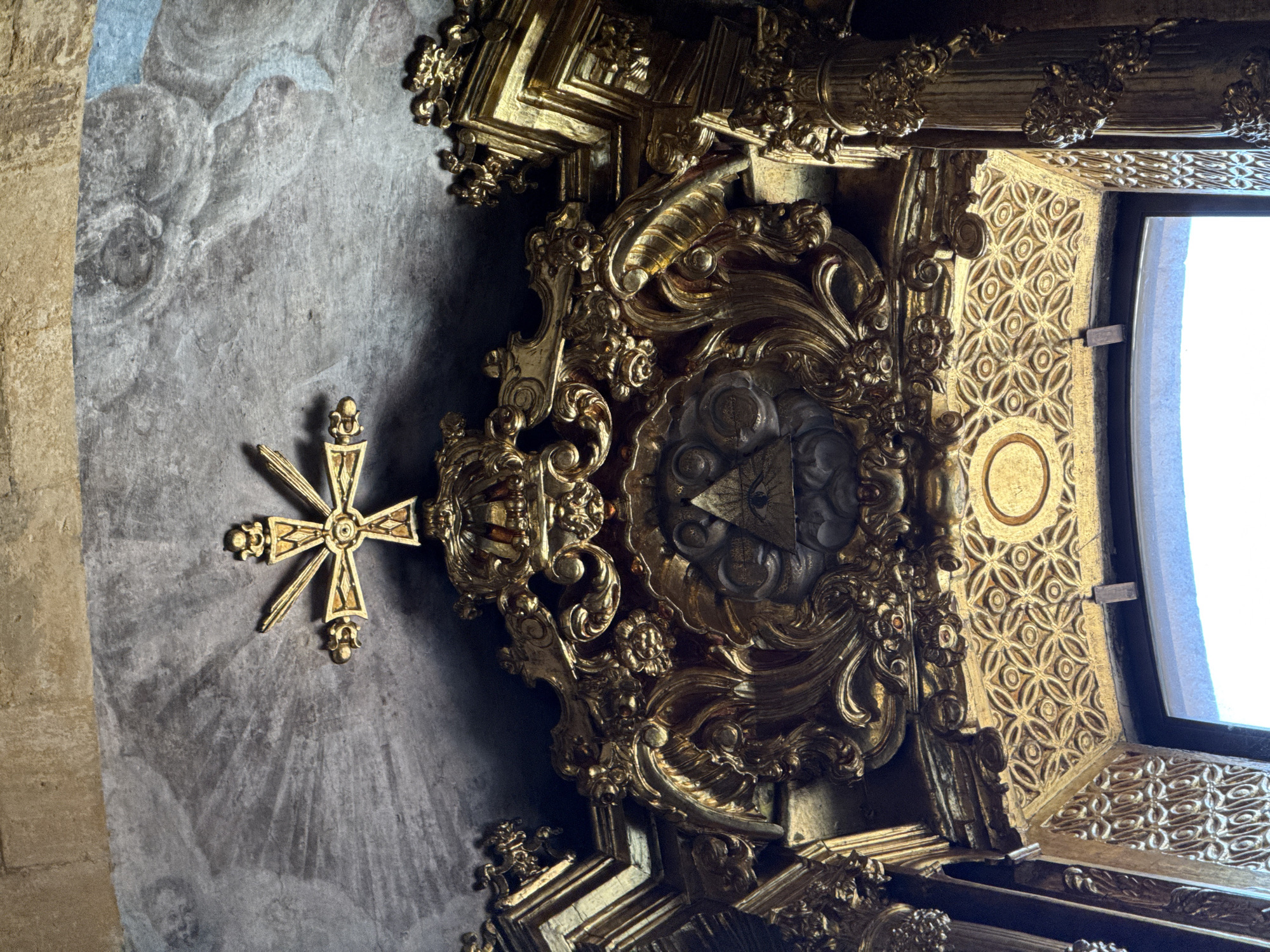 A low-angle shot shows a highly ornamented ceiling with a window in Unknown, Unknown. The ceiling is decorated with ornate gold carvings and moldings. A gold cross is affixed to the ceiling near a section painted with cloud imagery. A window is visible with a gold decorative lattice.