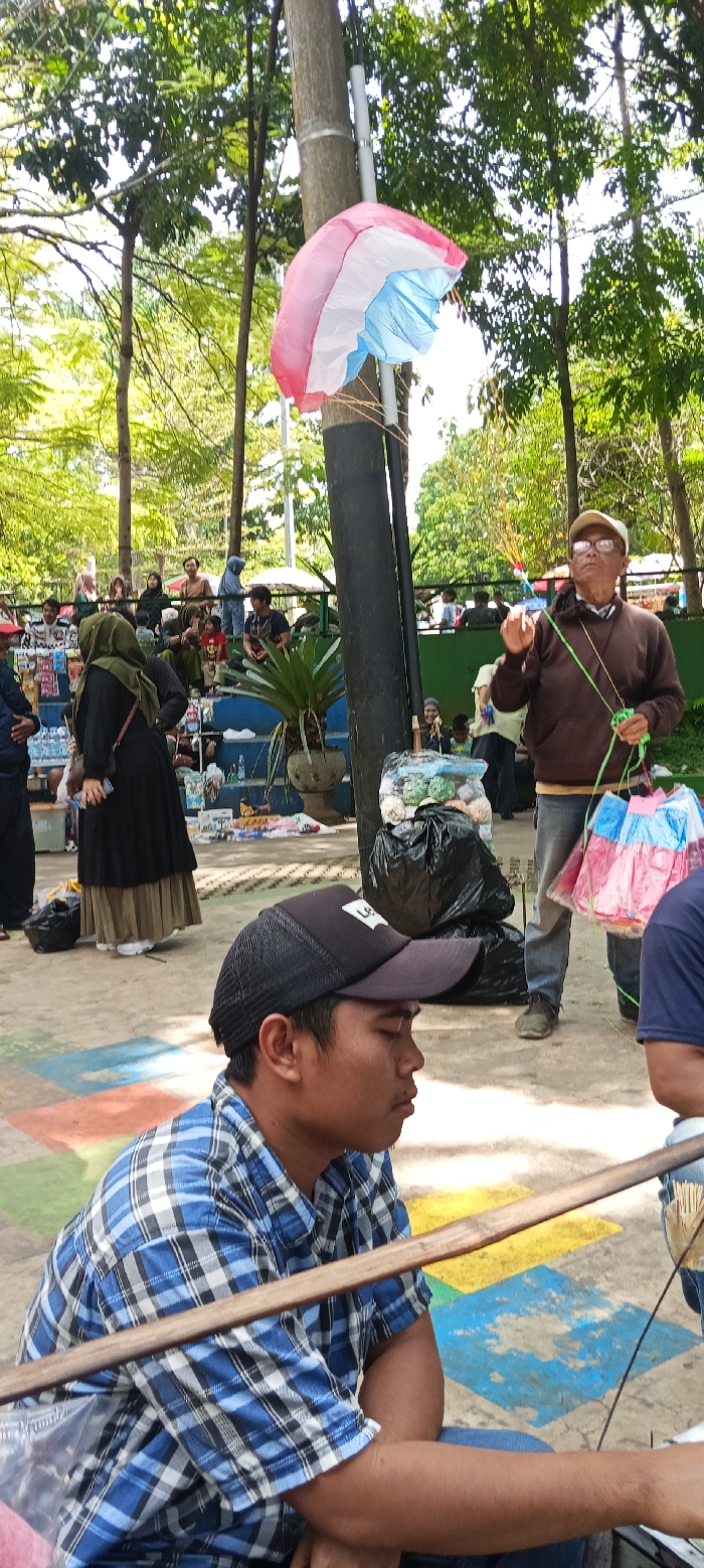 The scene takes place in a park in Soreang, Indonesia, located at latitude -7.023 and longitude 107.528. Several people are present, including a man wearing a brown shirt and a blue hat, holding a string attached to a pink and blue parachute.  A woman in a black and brown hijab is standing nearby. A man wearing a blue plaid shirt and a black baseball cap is sitting on the ground. He appears to be looking down, perhaps at the ground or an object in his hand. The park is covered in green trees. The ground is painted with a colorful pattern of red, yellow, blue, and green. There are black plastic bags filled with trash scattered around the park.  A white wooden fence can be seen in the background.