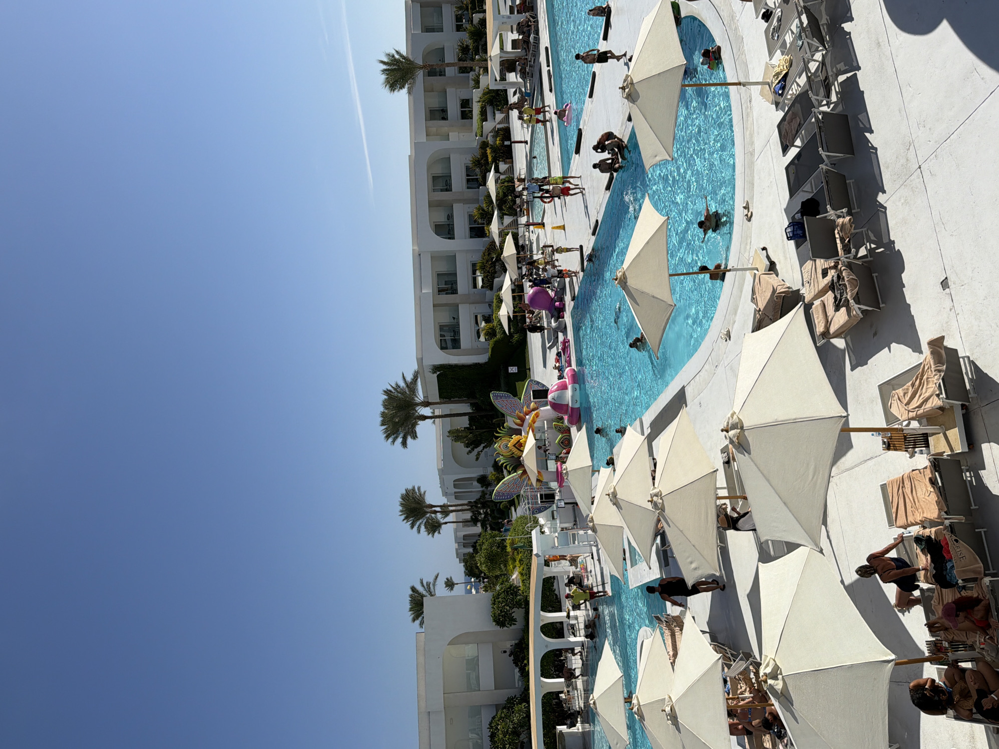 This is an aerial, daytime, eye-level shot of a resort pool area in Sharm El Sheikh, Egypt. The main focus is a large, bright blue, kidney-shaped swimming pool, filled with clear water and several people swimming and wading. Surrounding the pool is a paved area with numerous lounge chairs, many of which are occupied by people relaxing. Several large, beige beach umbrellas are deployed, providing shade.
In the background, white, modern resort buildings with multiple stories and arched doorways line the property. Lush green palm trees and other vegetation are scattered throughout the resort, suggesting a tropical or subtropical climate.
A prominent feature near the pool is a colorful, large inflatable butterfly structure, along with other inflatable toys, adding to the recreational atmosphere. There are also several yellow cones and a lifebuoy visible near the edge of the pool. People are seen in swimwear, enjoying the water and the sun. The sky is a clear, bright blue. The overall impression is a lively and relaxing resort environment.