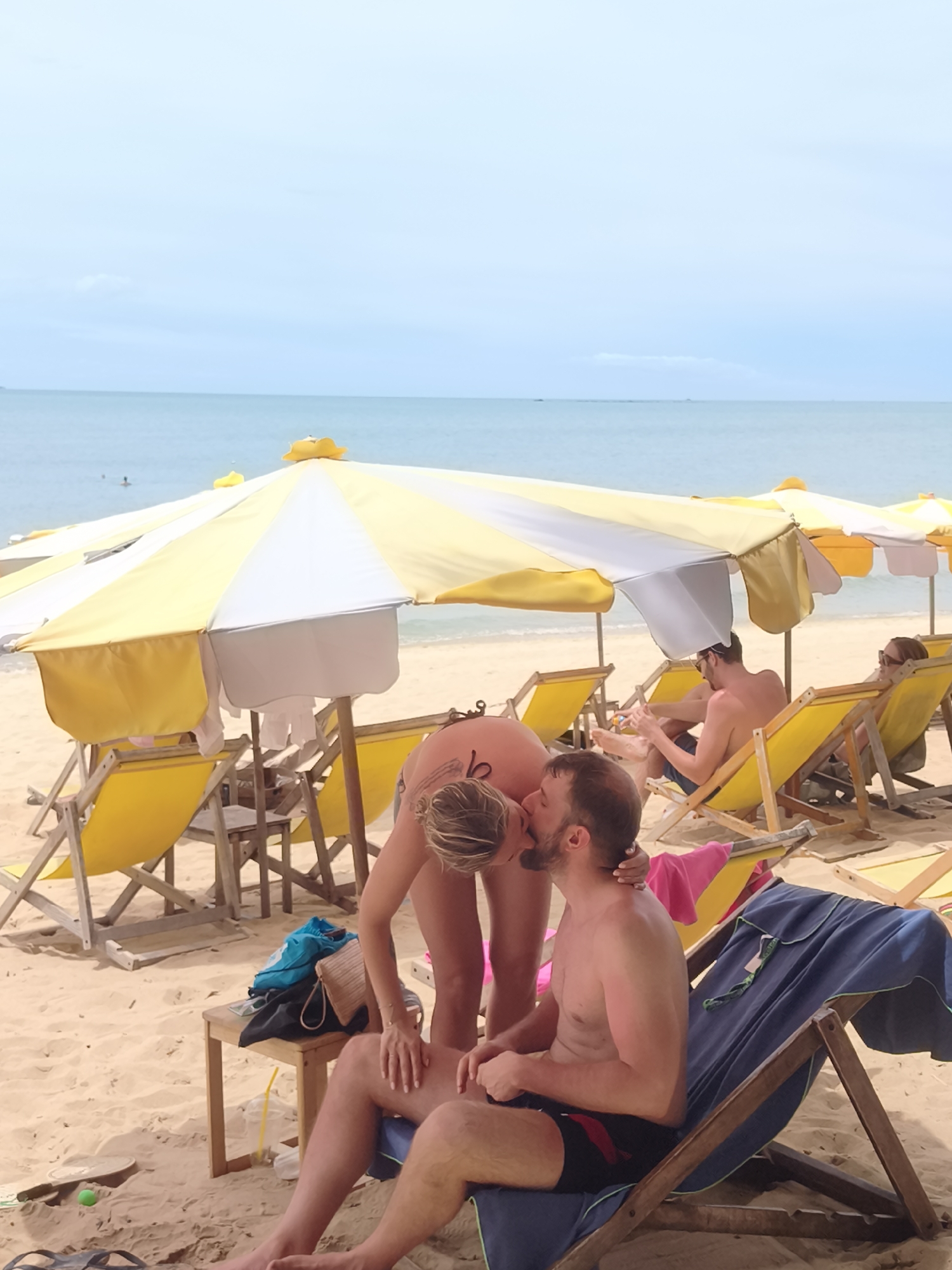 At Baan Mae Nam beach in Thailand, a couple is embracing and kissing on the sand, with one person kneeling and the other seated on a beach chair. The scene is set under yellow and white beach umbrellas, with several empty yellow lounge chairs in the background. The ocean is visible in the distance, under a pale sky. Other people can be seen relaxing on the beach further away. The sand is tan, and there are beach accessories like bags and towels scattered around.