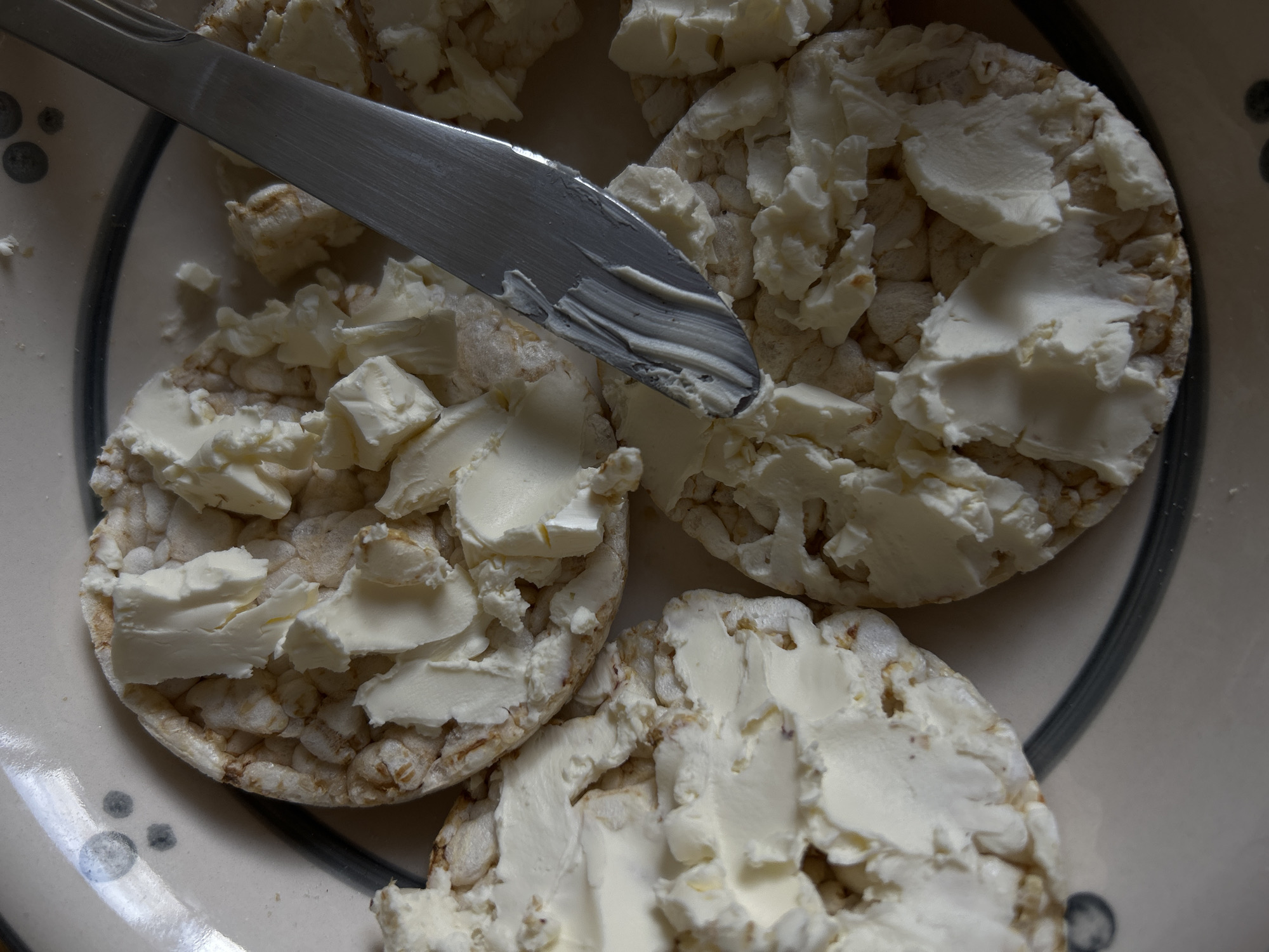 A close-up shot depicts a plate containing rice cakes topped with cream cheese. A metal butter knife spreads the cream cheese on one of the rice cakes. The plate has a light-colored background with dark gray decorative elements. The scene does not provide any location or information indicating it's in Sofia, Bulgaria.