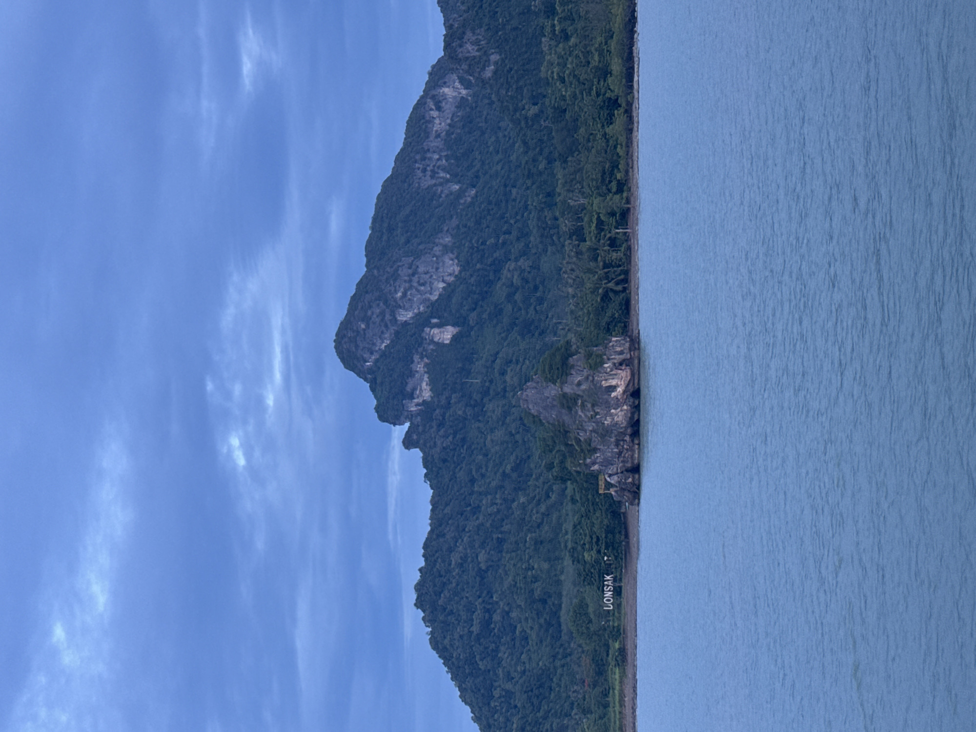 The image depicts a coastal scene with a large, lush green mountain rising from the sea. The mountain is covered in dense vegetation and shows some rocky outcrops. The sea is a calm, pale blue with ripples on its surface. The sky above is a clear, bright blue with some wispy white clouds. In the lower right portion of the mountain, white lettering spelling out "LOMSAK" is visible, suggesting a place name or sign. A small, yellow structure is also visible on the side of the mountain near the water's edge. The overall impression is of a serene, natural landscape, possibly taken during the daytime. The orientation of the image is vertical, with the mountain dominating the left side and the sea the right.