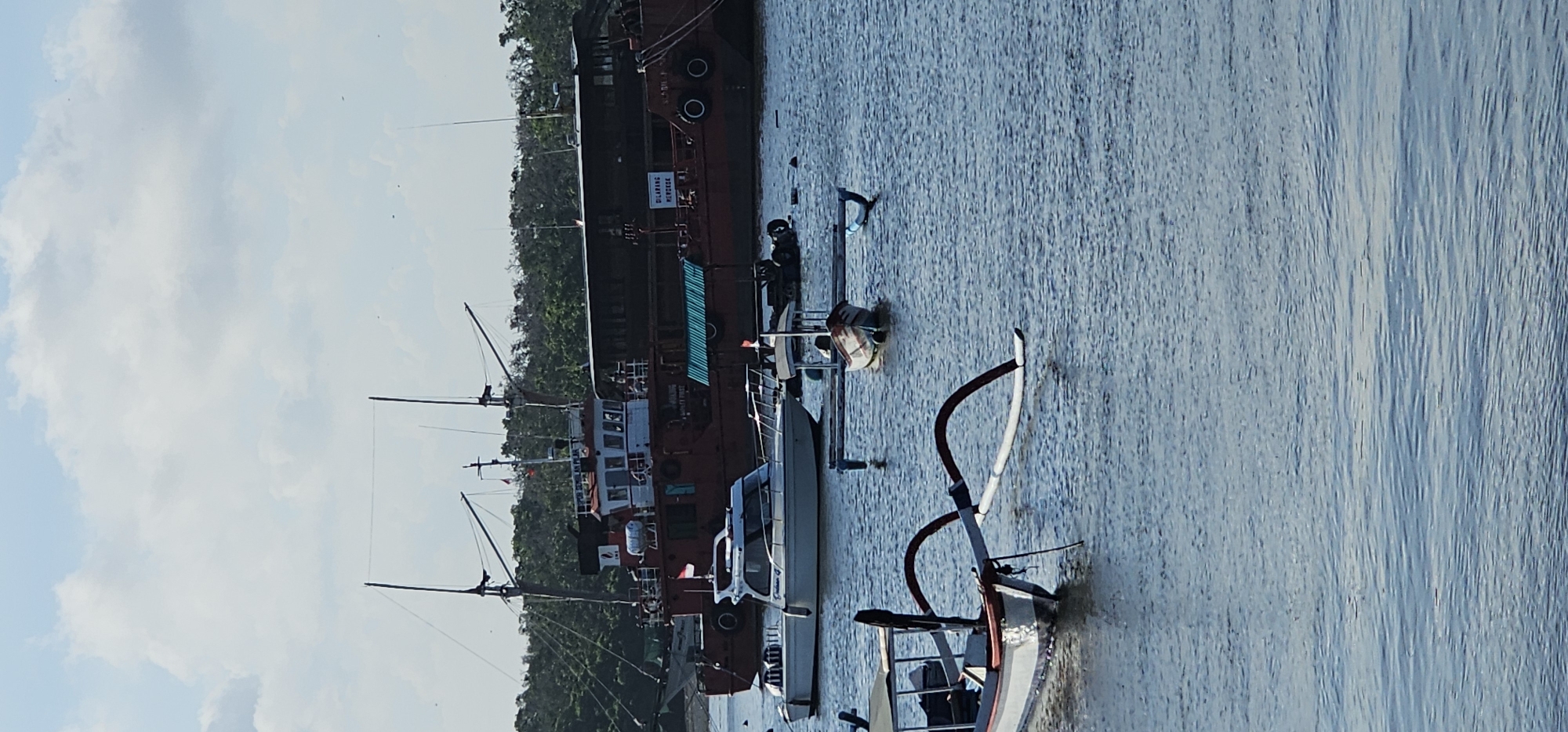 The scene shows a variety of vessels docked along a waterway in an unidentified location within Indonesia. A large, dark-hulled ship with multiple levels and visible equipment like antennas and signs is moored alongside smaller vessels, including speedboats and other watercraft. The waterway reflects the blue tones of the sky, which is partially obscured by trees lining the shore. A smaller boat with a distinctive curved frame sits in the foreground, adding a sense of depth to the composition. The background reveals lush green vegetation, possibly indicating a riverbank or coastal area.