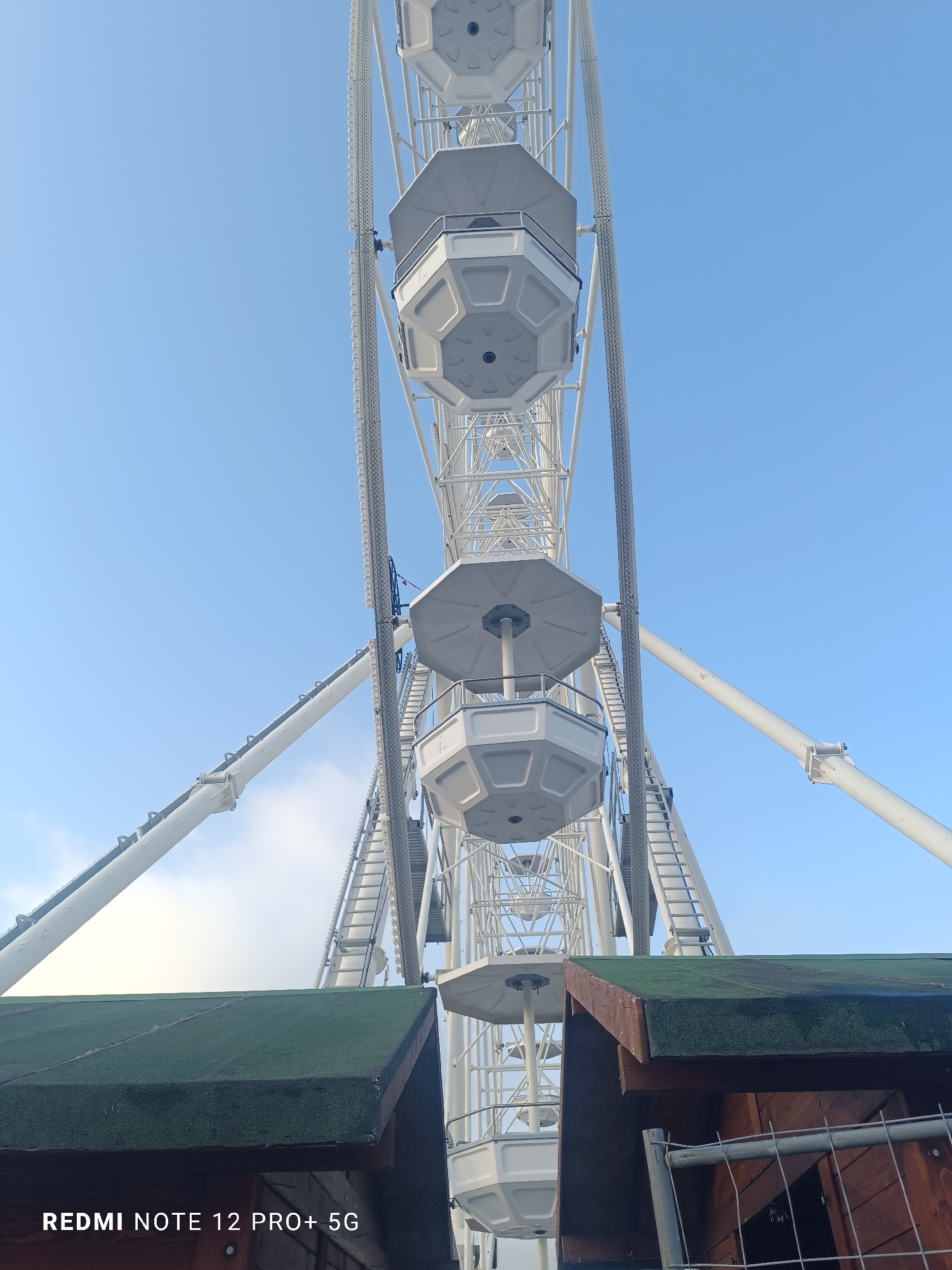 The image displays a large white Ferris wheel, viewed from a low angle looking upwards against a clear, bright blue sky. The intricate white metal framework of the Ferris wheel is prominent, with several enclosed gondolas (cabins) visible. These gondolas feature octagonal tops and bases, and are also white. The wheel's large support beams extend outwards, emphasizing its scale.
The sky is a brilliant blue, indicating a sunny day, likely morning or afternoon, with only a few faint white clouds visible towards the lower left horizon.
In the foreground, at the bottom of the image, the top portion of a small wooden structure is visible. It has a distinct dark green, textured roof and dark brown wooden siding. To the immediate right of this structure, a metal fence or railing is partially seen.
No people are visible, and no specific activity is taking place, but the scene clearly depicts an amusement park or fairground setting.
The text "REDMI NOTE 12 PRO+ 5G" is present as a watermark in the bottom left corner of the image. The location of this scene is Varna, Bulgaria.
