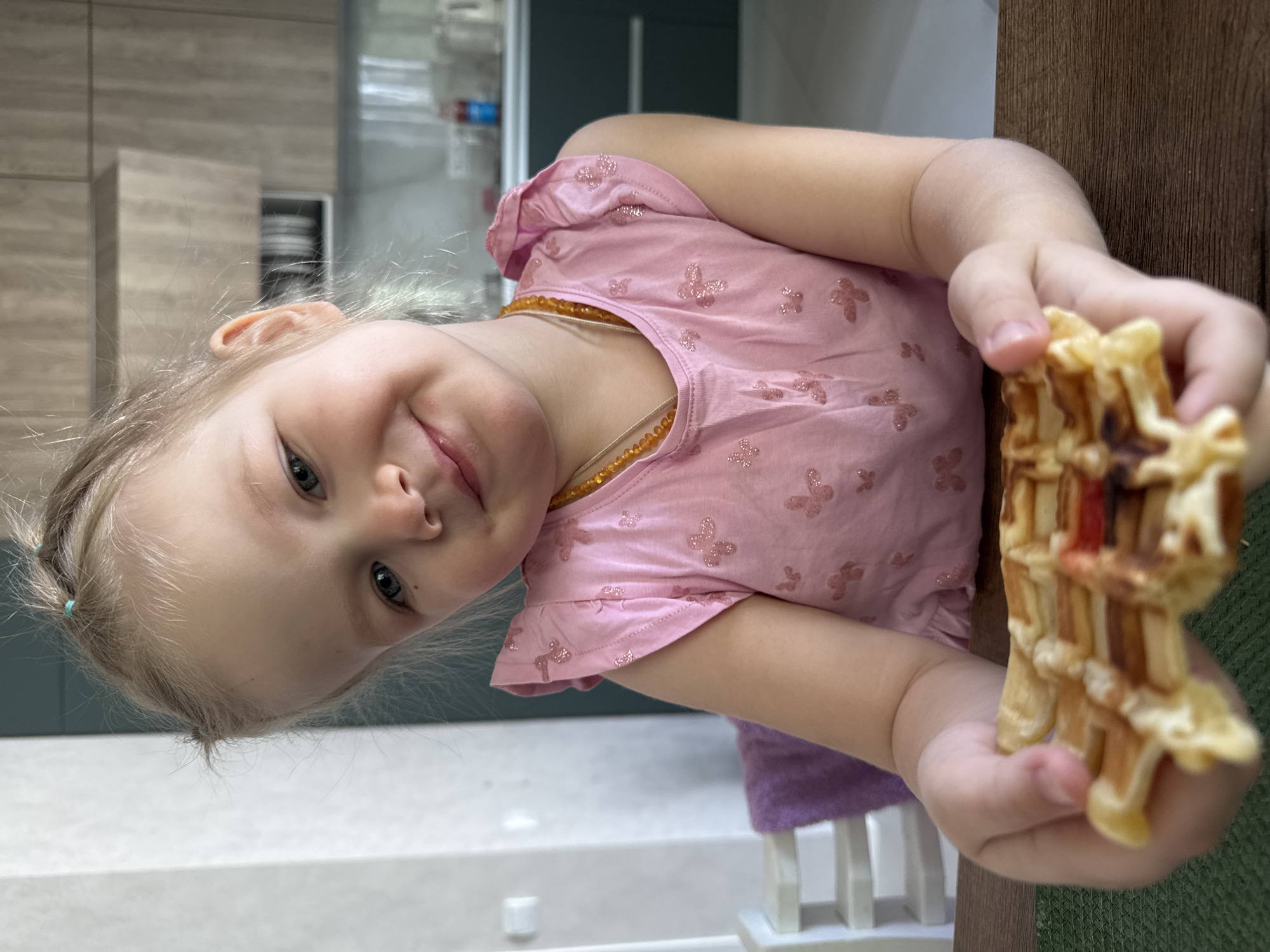 A young Caucasian girl with light blonde hair, styled in multiple small pigtails secured with light blue elastic bands, is seated at a dark wooden table, holding a golden-brown rectangular waffle with both hands. Her face, featuring blue eyes and a slight smile, is directed towards the camera. She wears a pink short-sleeved top with ruffled shoulder details and glittery butterfly patterns, along with an amber bead necklace. A green placemat is partially visible beneath her hands. To her left, part of a white chair with a purple cushion is discernible. The background reveals a modern kitchen interior with light wood-grain upper cabinets, an integrated extractor hood, and dark grey/blue lower cabinets. A white rectangular wall fixture is present on the light gray wall to the left, and various kitchen items are visible on the counter. The scene is located in Gornyi (Dvurechenskoe S/P), Russia.