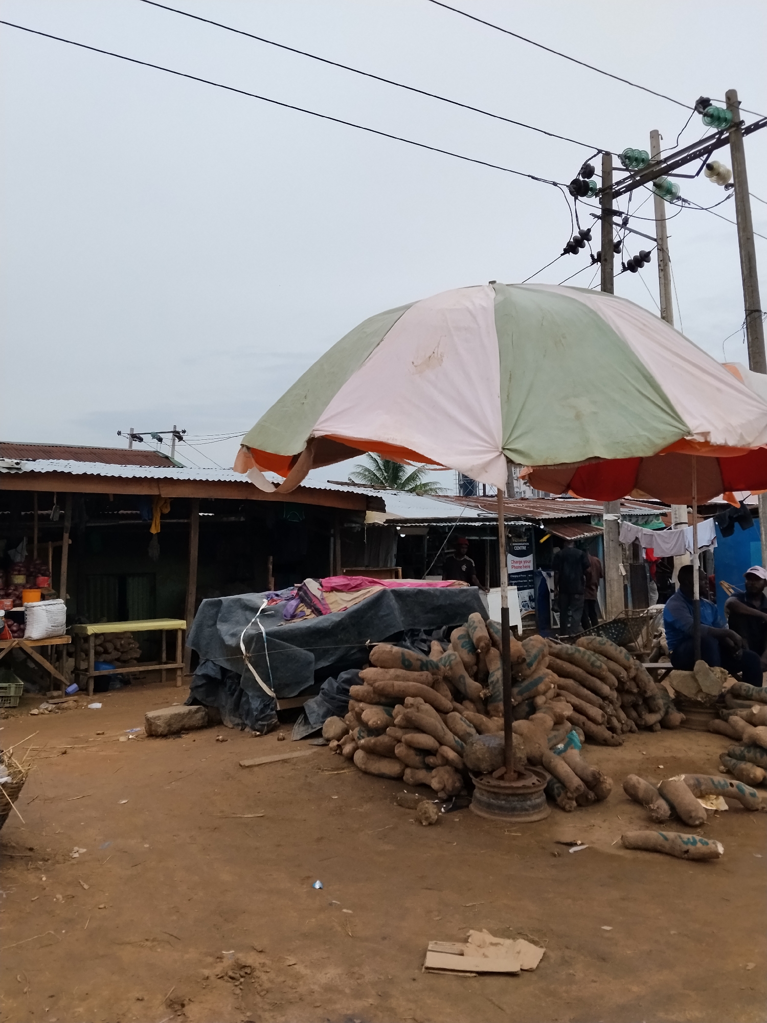 The image depicts a marketplace in Ura Nta Umuarandu, Nigeria. Several piles of yams are prominently displayed under a large, multi-colored umbrella. In the background, market stalls with corrugated metal roofs are visible, and people are present, some seated and others walking. Power lines and utility poles are overhead. The ground is dirt and scattered with debris.