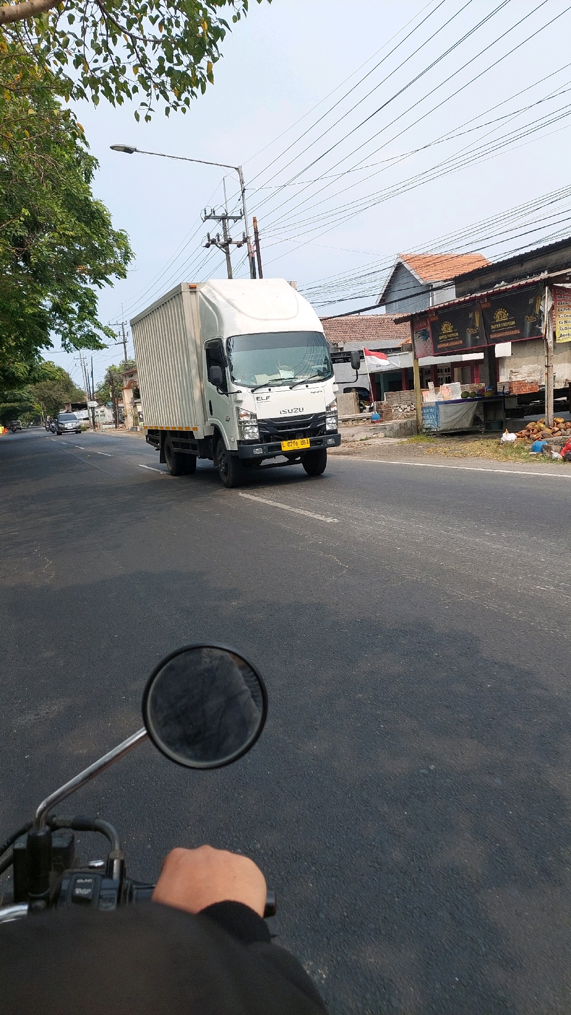 An eye-level, wide shot captures a white Isuzu Elf truck driving down a road in Ketimang, Indonesia. The truck, with a large white cargo container, is in motion on the asphalt road. In the foreground, a motorcycle rider's left hand is visible on the handlebars, and a portion of the rider's left shoulder is also in view. The rider's perspective is implied by the presence of the motorcycle's side mirror, reflecting a blurred image. On the right side of the road, there are several small stalls with awnings and goods displayed, including coconuts and other produce. Trees with green foliage line the left side of the road, and utility poles with numerous wires are visible in the background. The sky is clear and light blue.